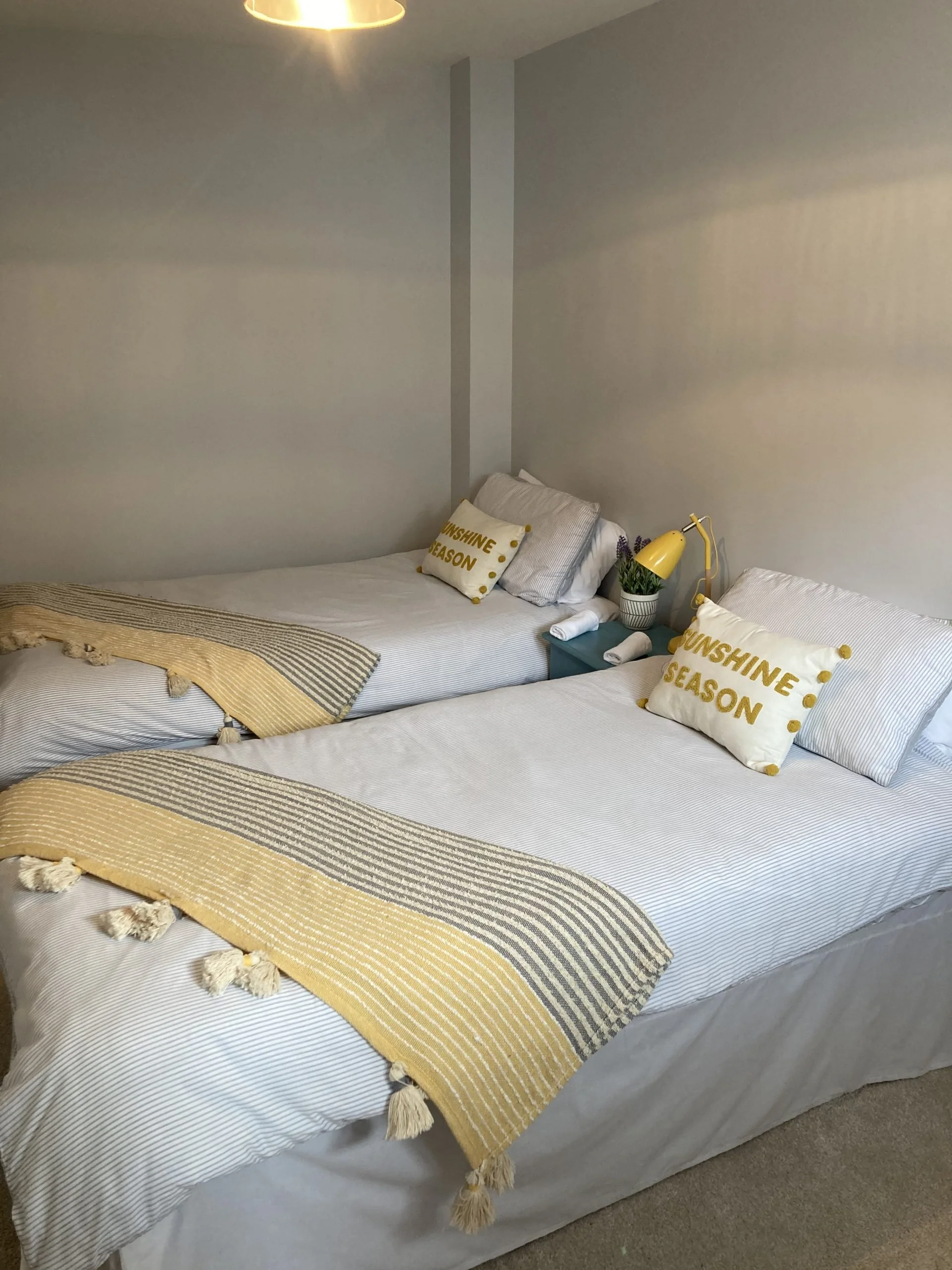 Two neatly made beds with white sheets and yellow and gray striped throws, each with pillows reading 'Sunshine Season.' A small teal nightstand between the beds holds a plant, a yellow desk lamp, and a couple of rolled towels. The room has light gray
