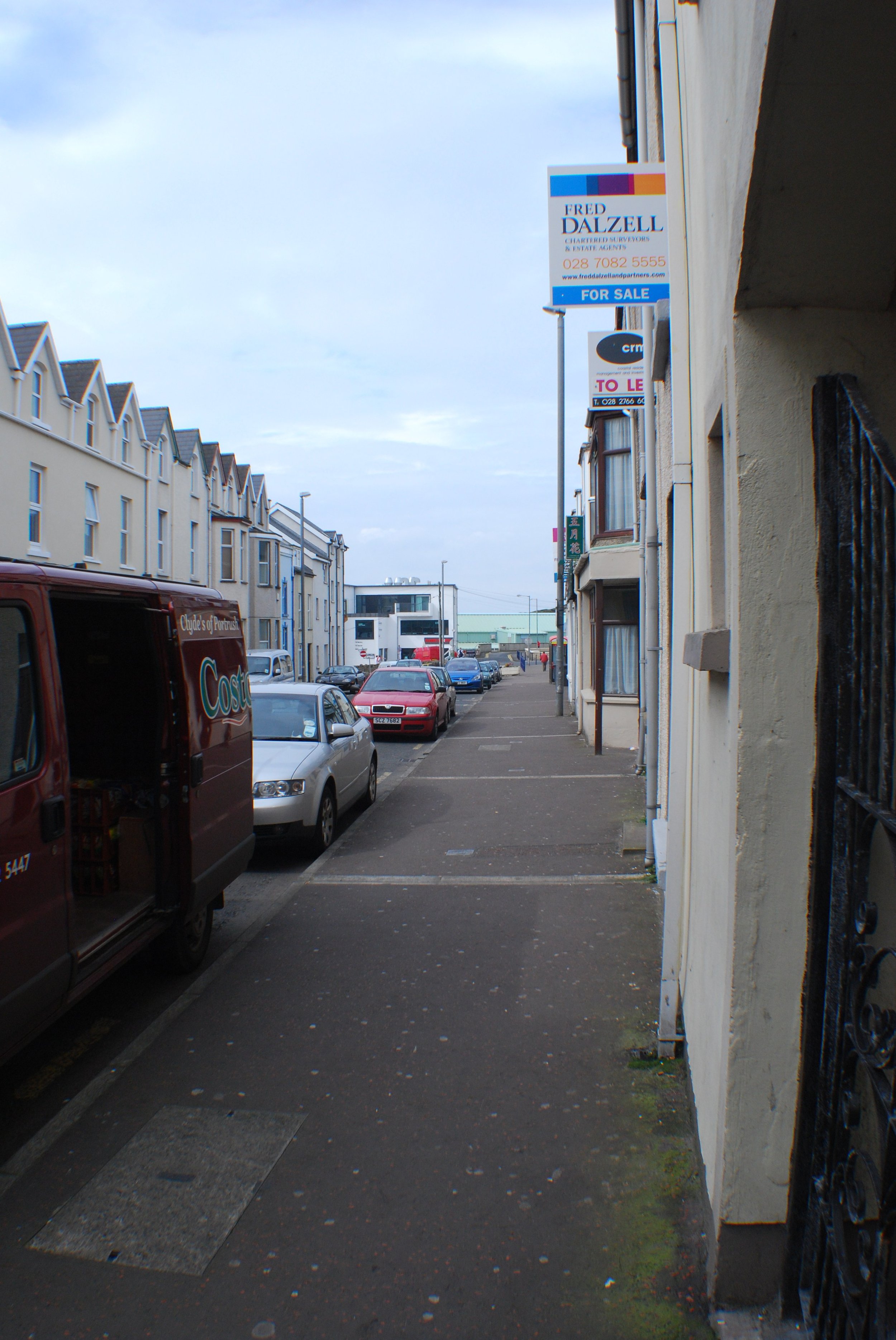 Empty sidewalk with parked cars along a row of buildings, with a 