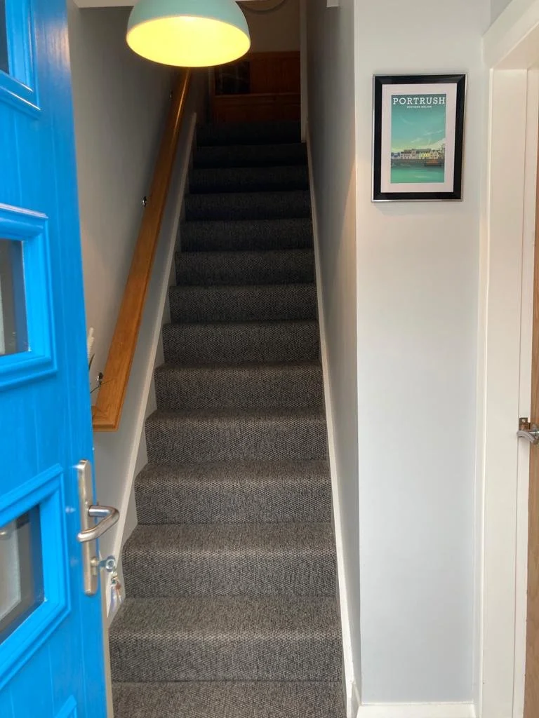 Indoor staircase with dark gray carpet, wooden handrail on the left, white walls, and a framed poster of Port Rush on the right wall, partial view of a blue door and a ceiling light.