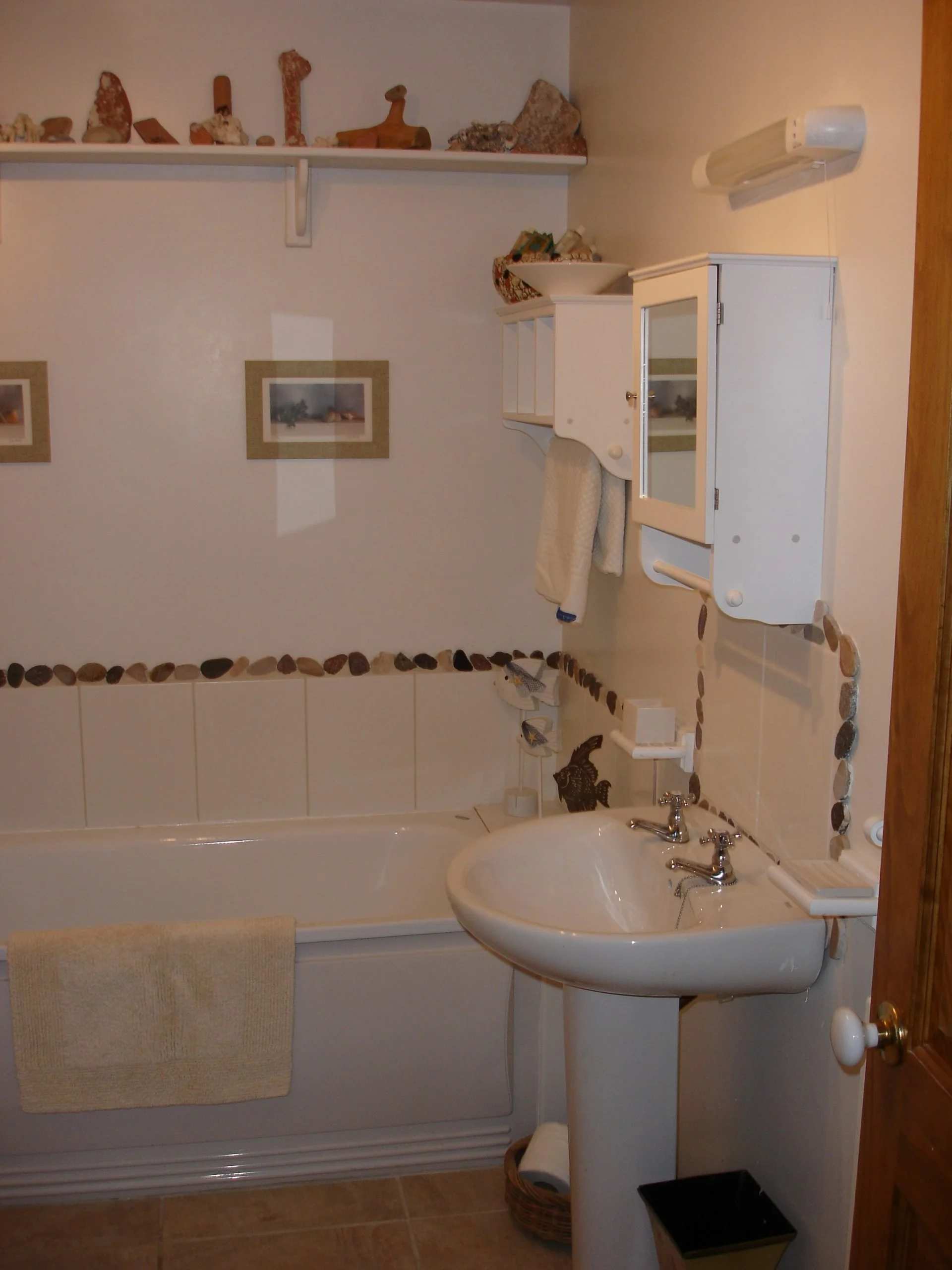 A small bathroom featuring a bathtub, a pedestal sink, white wall-mounted cabinets, a towel hanging on a rack, decorative river stones accenting the tile border, and framed pictures on the wall.