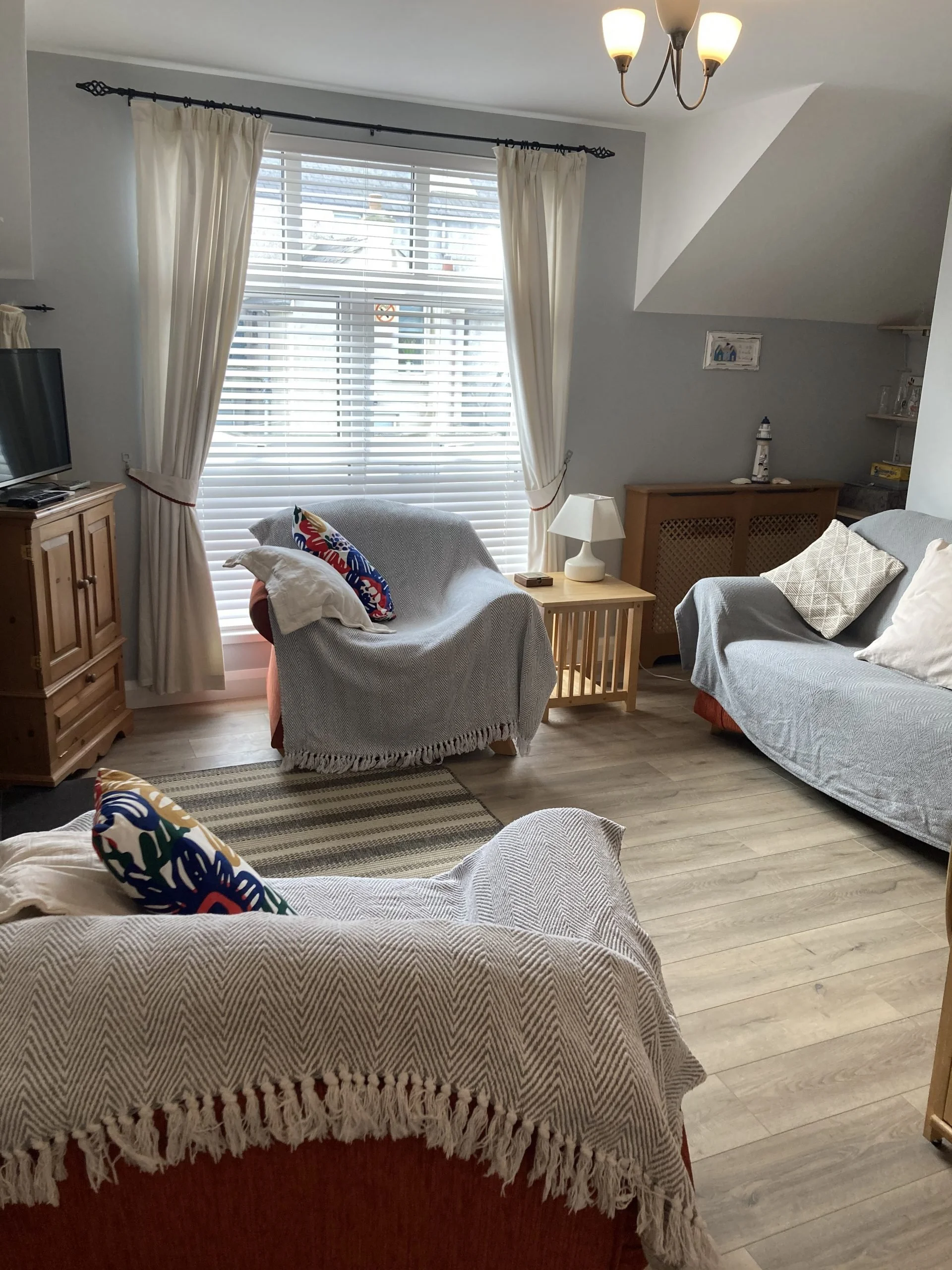 A cozy living room with a window covered by white blinds and cream curtains. There are three sofas covered with light-colored throws, each with decorative pillows. A wooden side table with a white lamp sits between two sofas, and a wooden cabinet wit