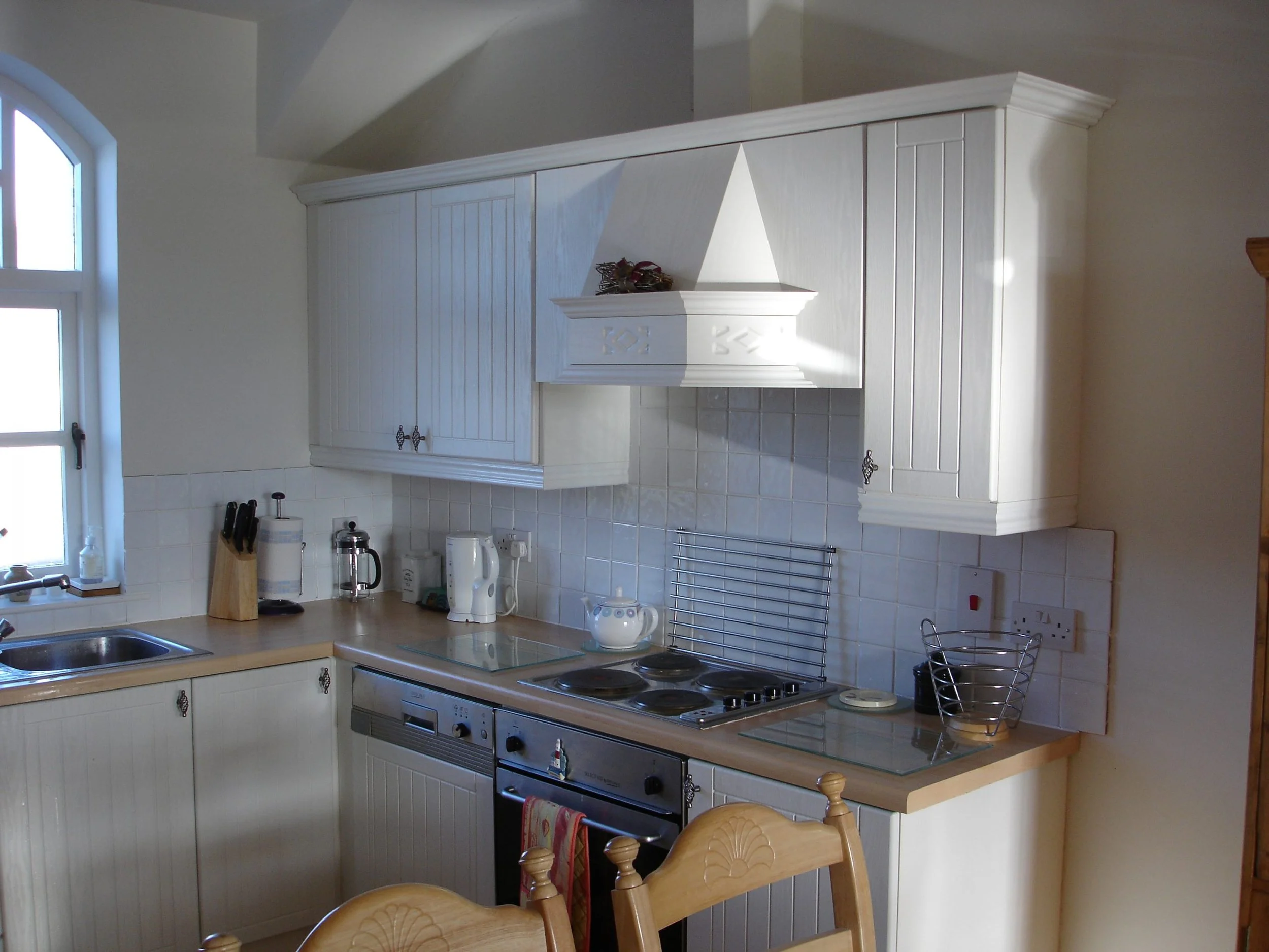 Kitchen with white cabinets, a stovetop, and a countertop with various kitchen appliances, a window on the left, and a wooden table with chairs in front.