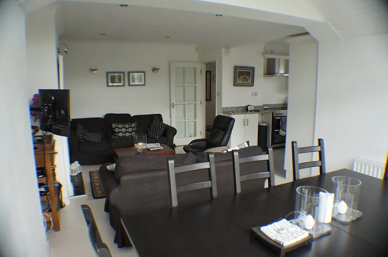Living room and dining area with black couches, a black office chair, a TV on a stand, a dining table with chairs, and a kitchen in the background.