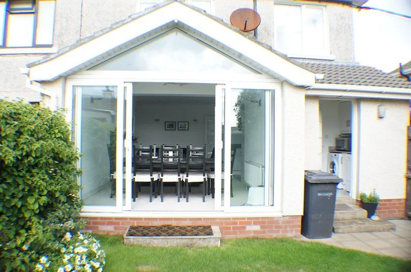 Backyard view of a house with a sunroom, interior dining table and chairs, and a side area with washing machine and microwave.