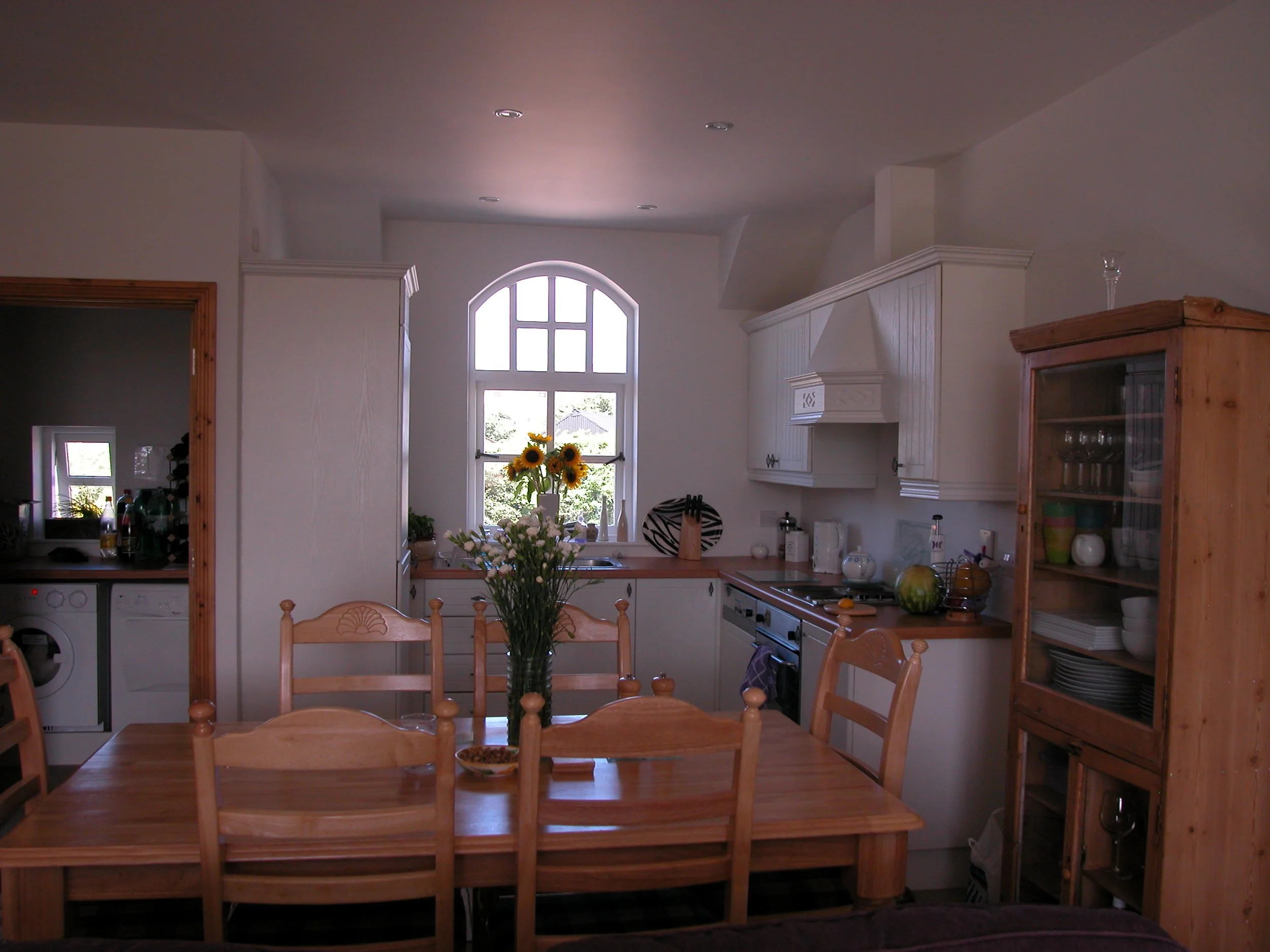 Kitchen with wooden dining table, sunflower vase, window with daylight, white cabinets, kitchen appliances, and glass cabinet with dishes.