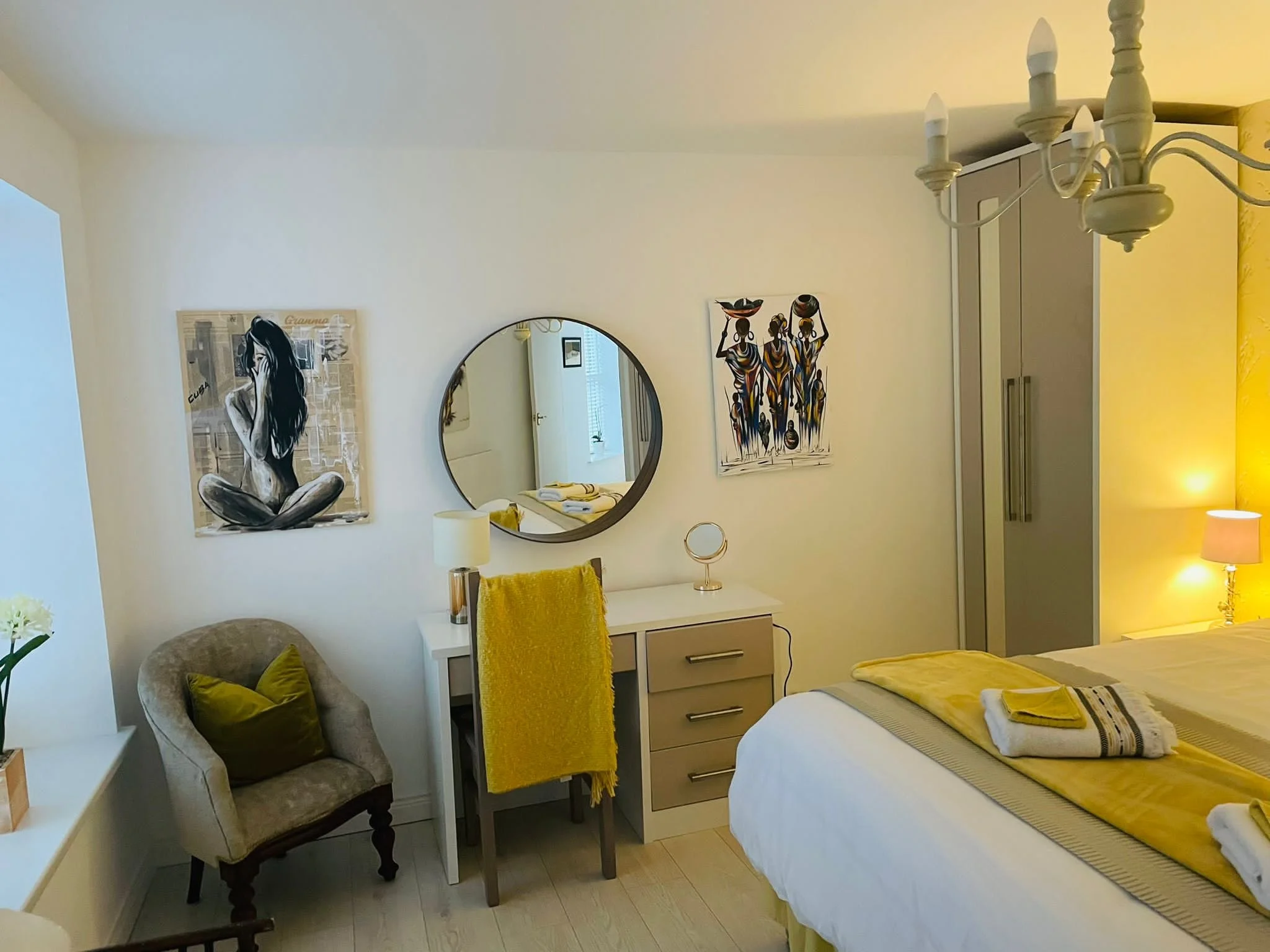 A bedroom with white walls, a bed with yellow and white bedding, a bedside lamp, a wardrobe, a white dresser with a small mirror, decorative art on the wall, a beige armchair with a green pillow, a round wall mirror, and a chandelier.