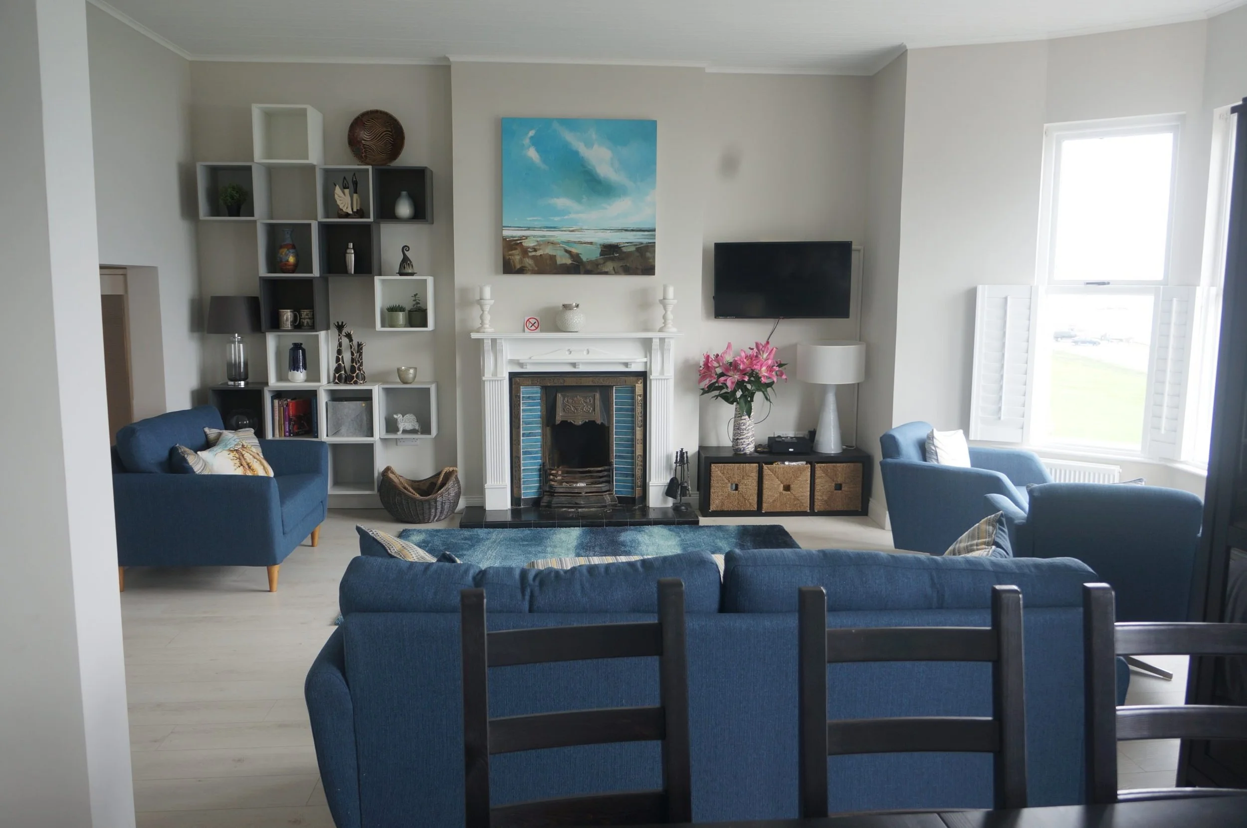 Living room with blue sofas, a patterned area rug, a fireplace with a painting above, a flat-screen TV, a side table with pink flowers, a window with open shutters, and decorative shelves with various items.