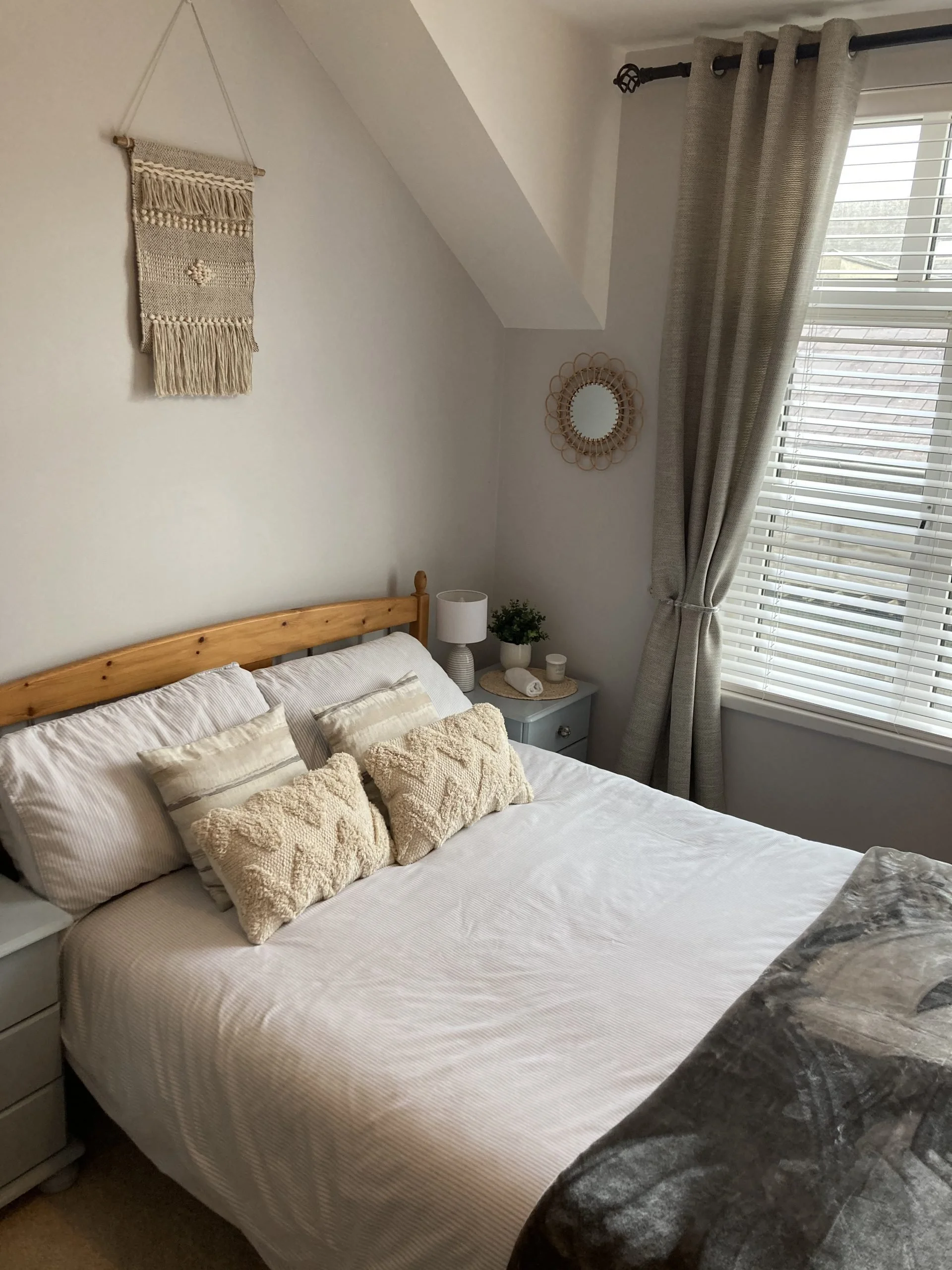 A cozy bedroom with a wooden bed frame, beige and cream pillows, a white bedsheet, a nightstand with a lamp, a plant, and decorative items, next to a window with gray curtains and white blinds. A wall hanging and a small round mirror are on the wall.