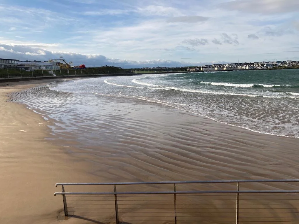 A sandy beach with gentle waves washing ashore, a metal railing in the foreground, and buildings along the shoreline under a partly cloudy sky.