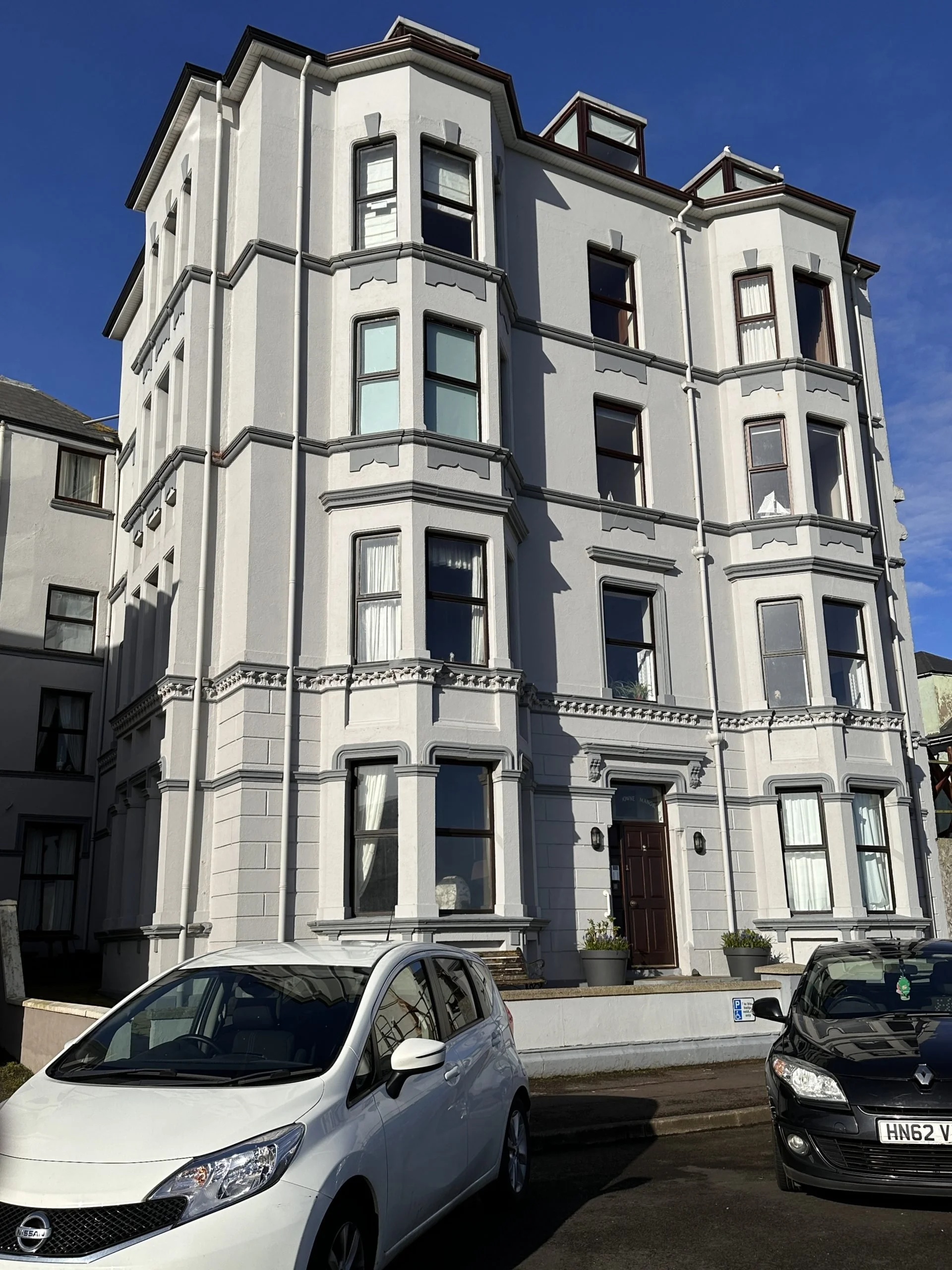 A white multi-story Victorian-style building with bay windows and decorative trim, situated on a street with parked cars, including a white Nissan and a black Renault, under a clear blue sky.