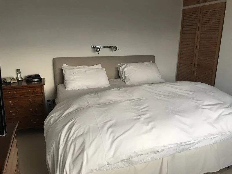 A neatly made hotel bed with white linens, two pillows, a wooden nightstand with a phone and alarm clock, and a wooden closet in the background.