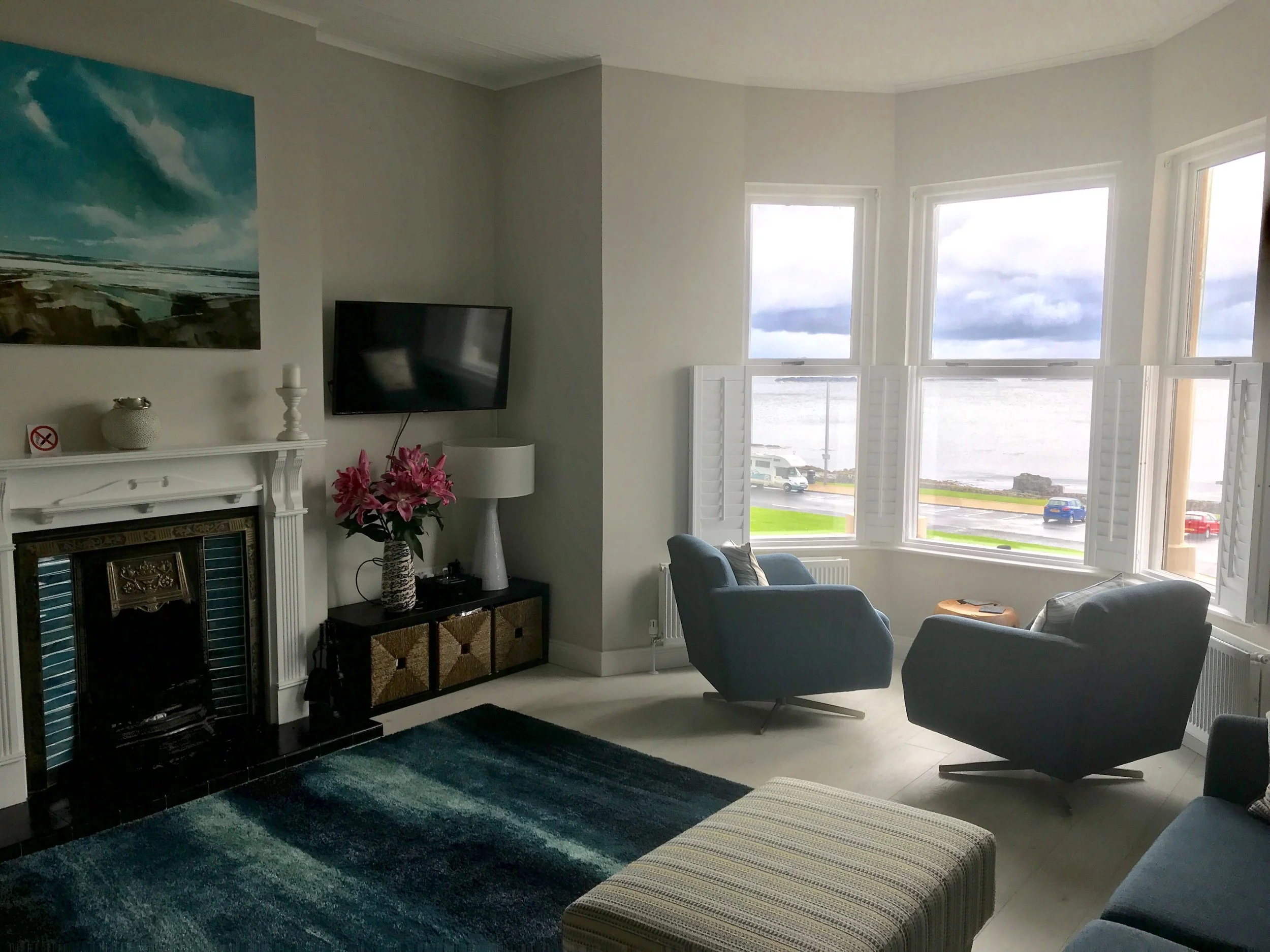 Living room with a fireplace, a flat-screen TV, floral lamp, blue armchairs, and large windows showing a view of a cloudy sky over a body of water and parked cars.