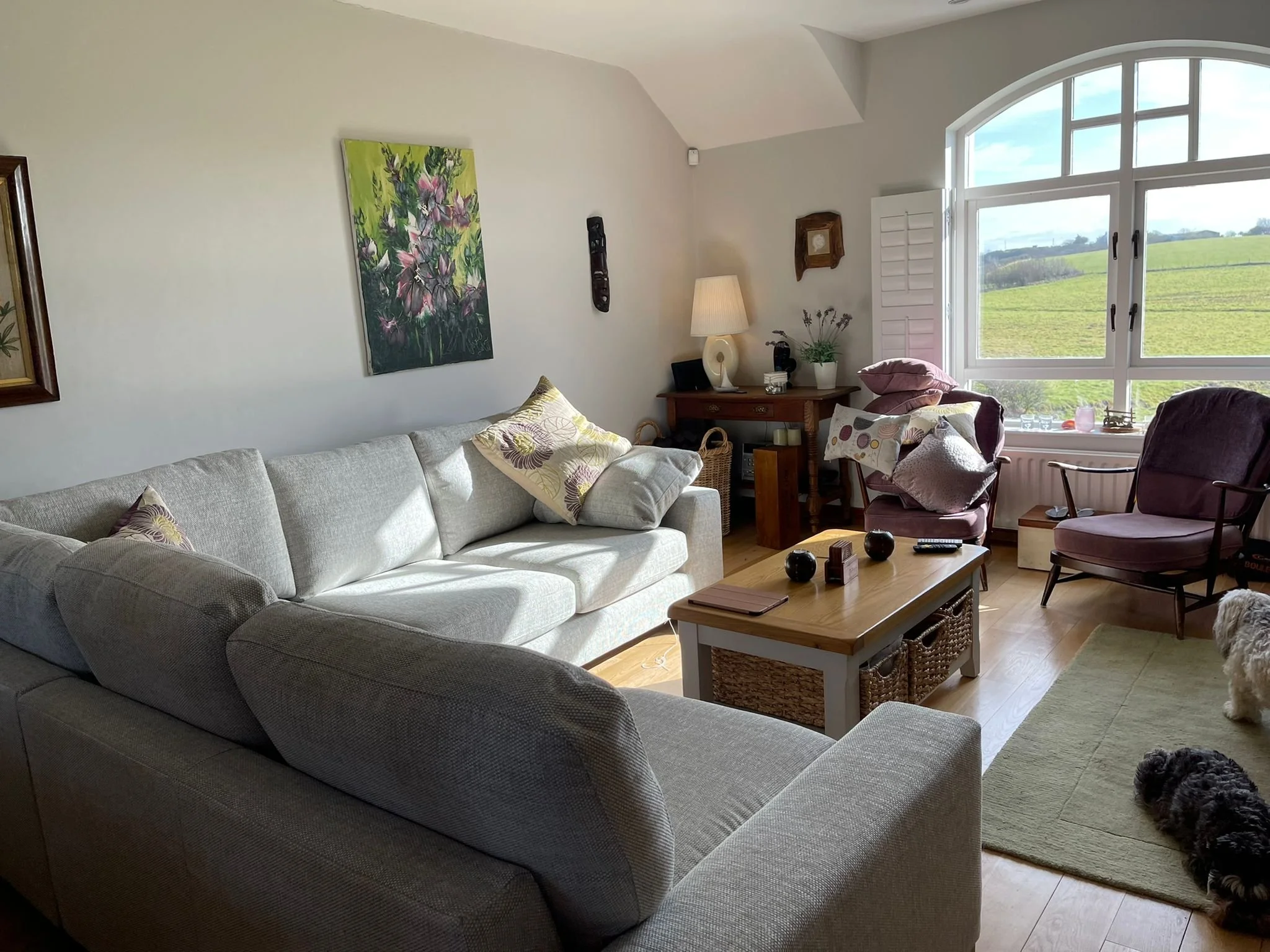 Living room with a large light-colored sofa, a coffee table, two purple armchairs with pillows, a window showing green fields outside, a lamp, and decorative items.