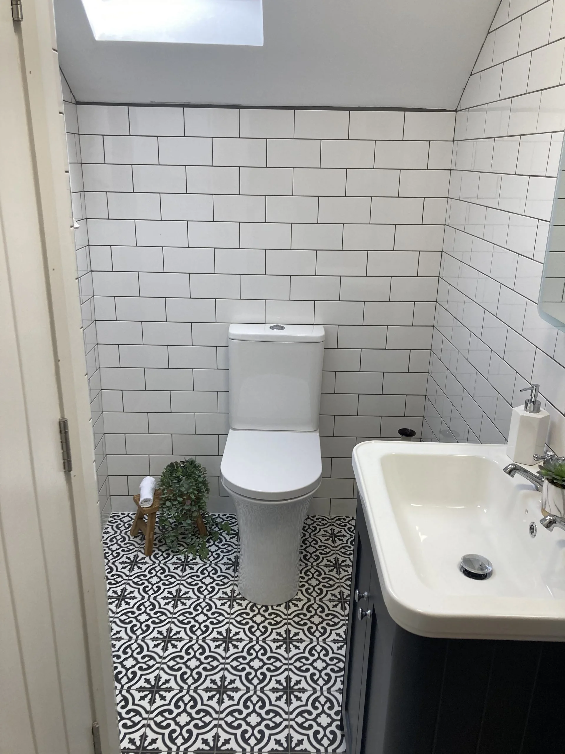 A small bathroom with white subway tile walls, patterned black and white floor tiles, a white toilet, a small sink with a cabinet below, a mirror, a soap dispenser, a small plant, and a towel on a wooden stand next to a green plant.
