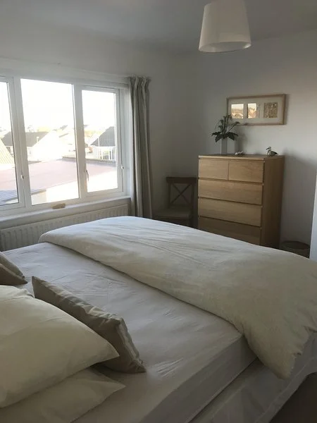 A bedroom with a large window showing rooftops outside, a bed with white bedding and pillows, a wooden dresser with a plant and a picture frame on top, a chair next to the dresser, and a ceiling light fixture.
