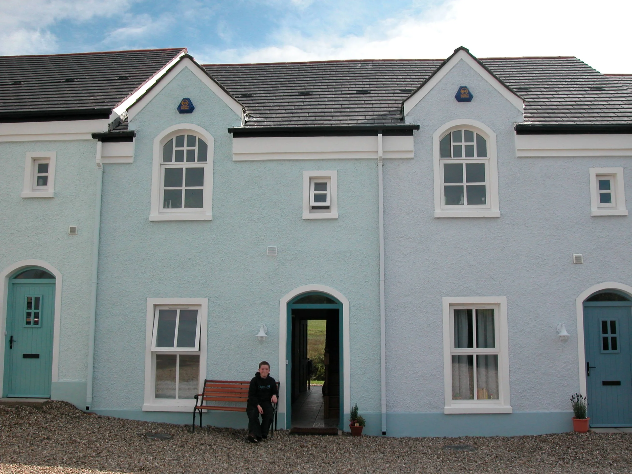 A boy standing outside a pastel blue and white two-story house with arched windows and doors, gravel ground, and potted plants.