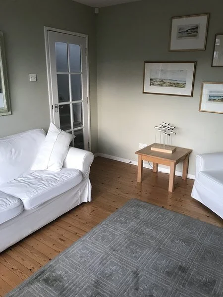 Living room with two white sofas, a small wooden table, framed pictures on the wall, a gray area rug, and a glass-paneled door.