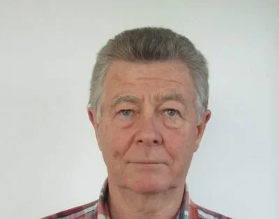 A close-up portrait of an older man with gray hair, blue eyes, and a serious expression, wearing a plaid shirt against a plain light background.
