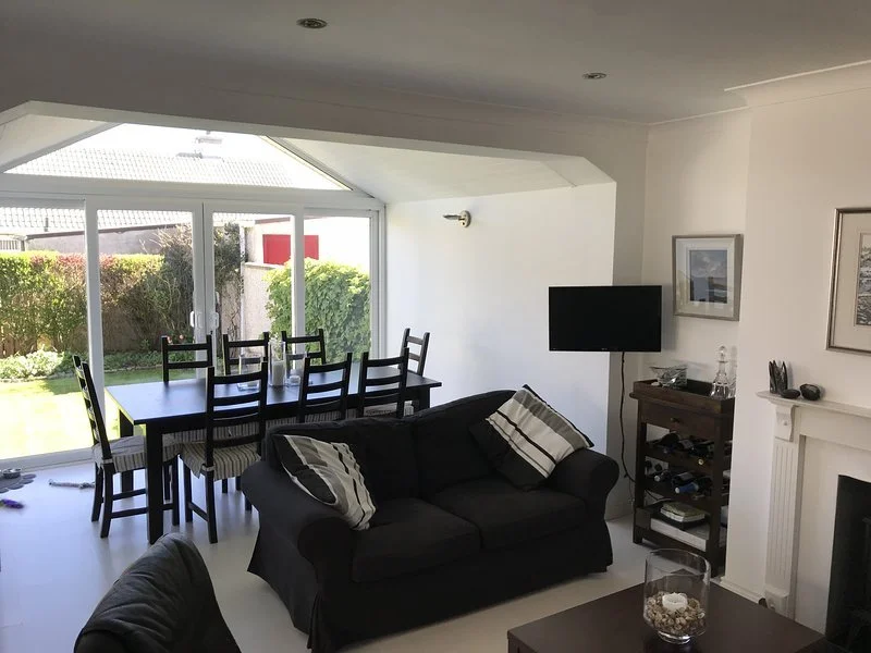 A living room with a black sofa, striped pillows, a small wooden cabinet, and a wall-mounted flat-screen TV. Adjacent to the living area, a dining table with eight chairs is positioned near large windows and glass sliding doors looking out to a garde