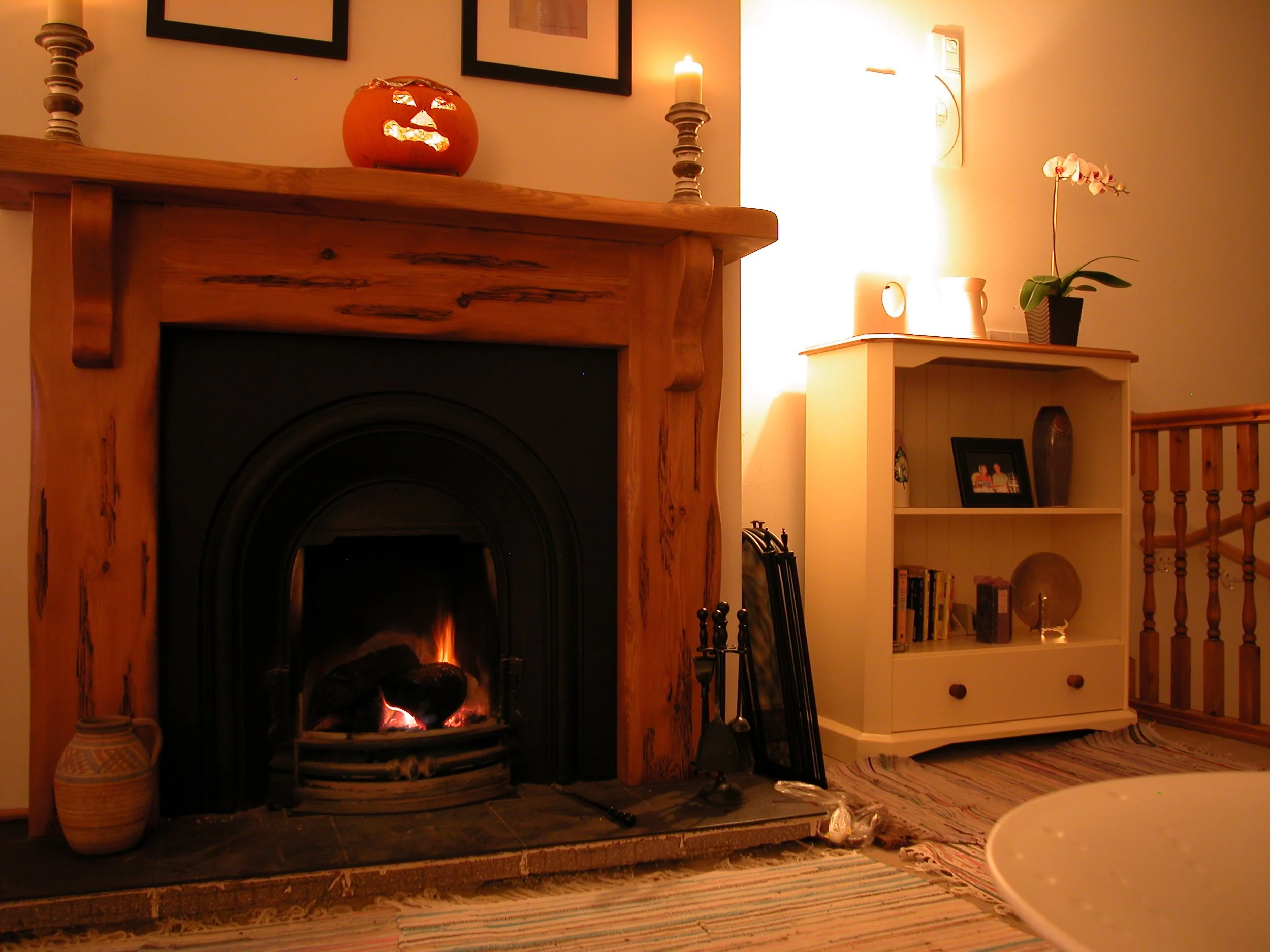 A cozy living room corner with a lit fireplace, a small wooden shelf with books and decorative items, a vase of orchids on the shelf, a lit candle, and a pumpkin with carved faces on the mantel.