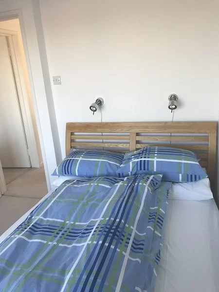 A neatly made bed with a wooden headboard, blue plaid bedding, and two matching pillows, in a bright bedroom with a white wall and an open door leading to another room.