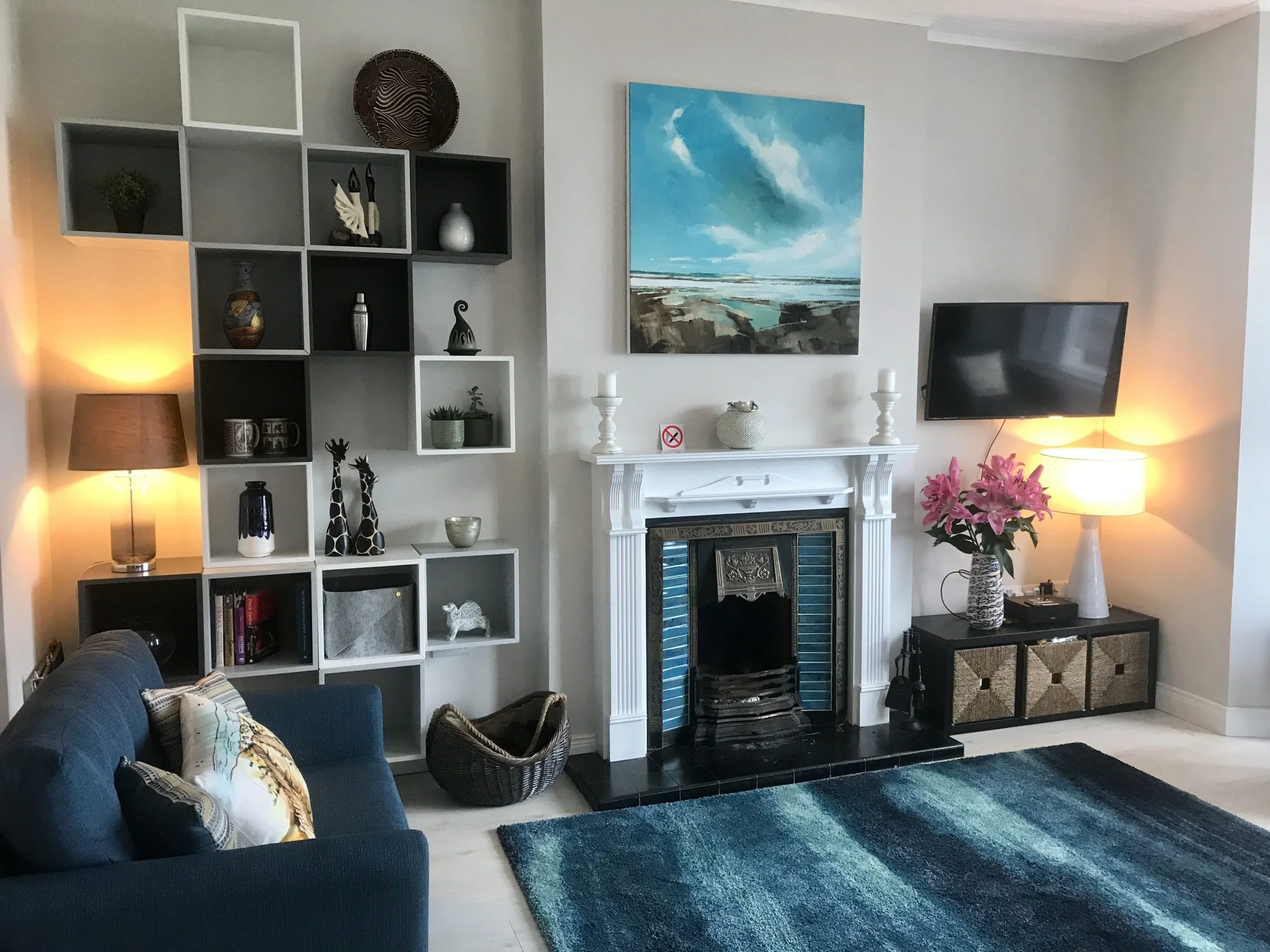 Living room with white walls, decorated with a large painting of a seascape above a white fireplace. To the right, a flat-screen TV on a black cabinet with pink lilies and a lamp. To the left, a blue sofa, a colorful rug, a white shelving unit with v