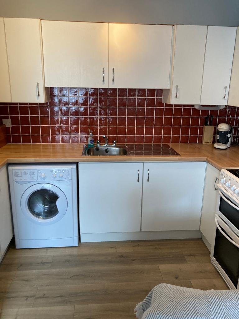 Kitchen with white cabinets, red tiled backsplash, wooden countertop, front-loading washing machine, sink, coffee maker, and stove.