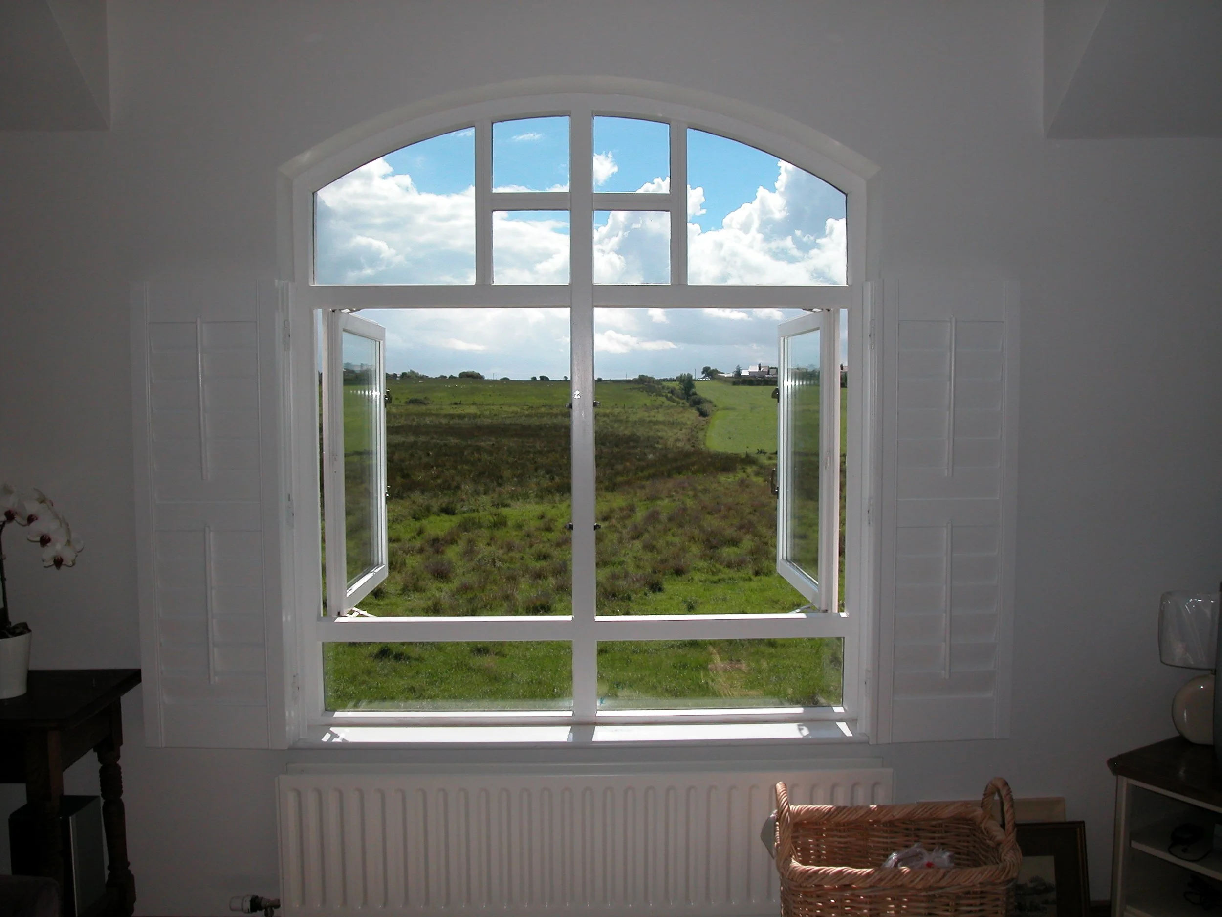 View through a large, arched window with open shutters, showing a green field and a partly cloudy sky outside.