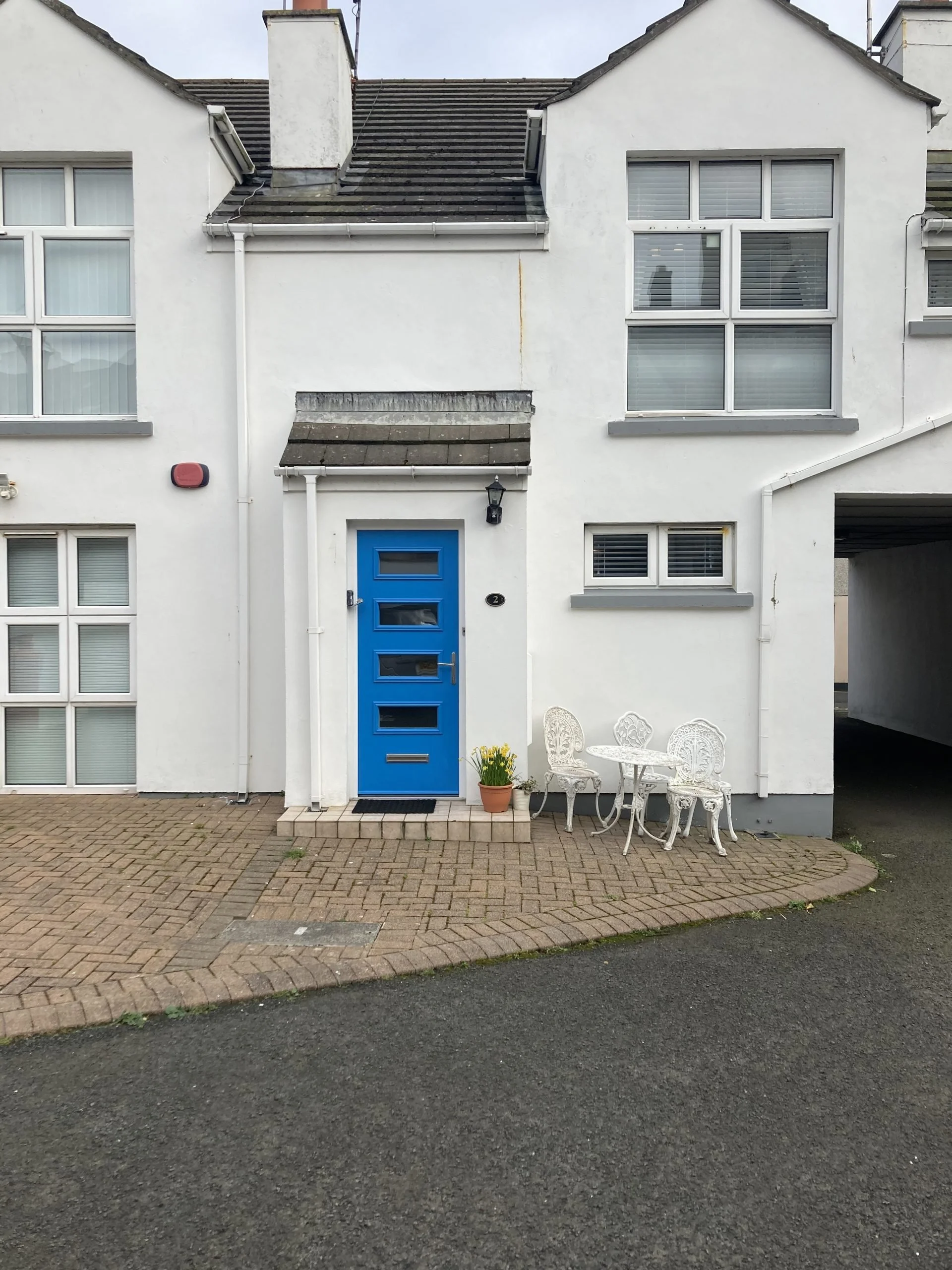 White house with blue front door, white outdoor table and two chairs, potted yellow flowers, and multiple windows.