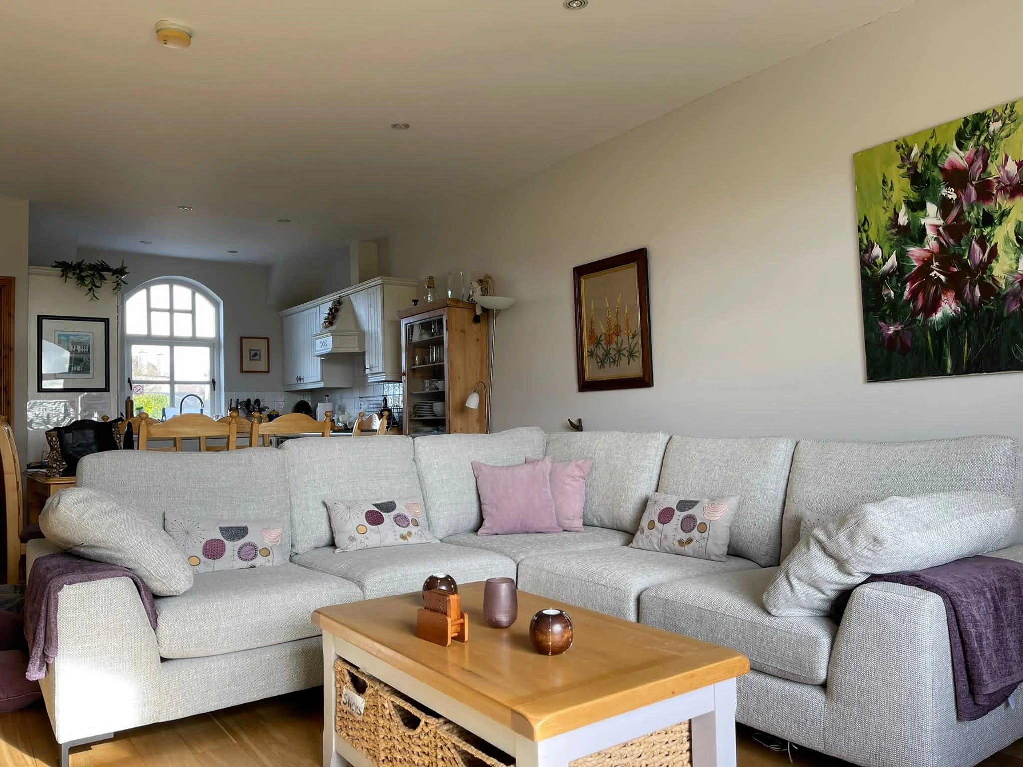 A cozy living room featuring a large, light-colored sectional sofa with pink and patterned throw pillows. There are decorative items on the coffee table. The background reveals a bright kitchen area with a wooden dining table, a window letting in nat