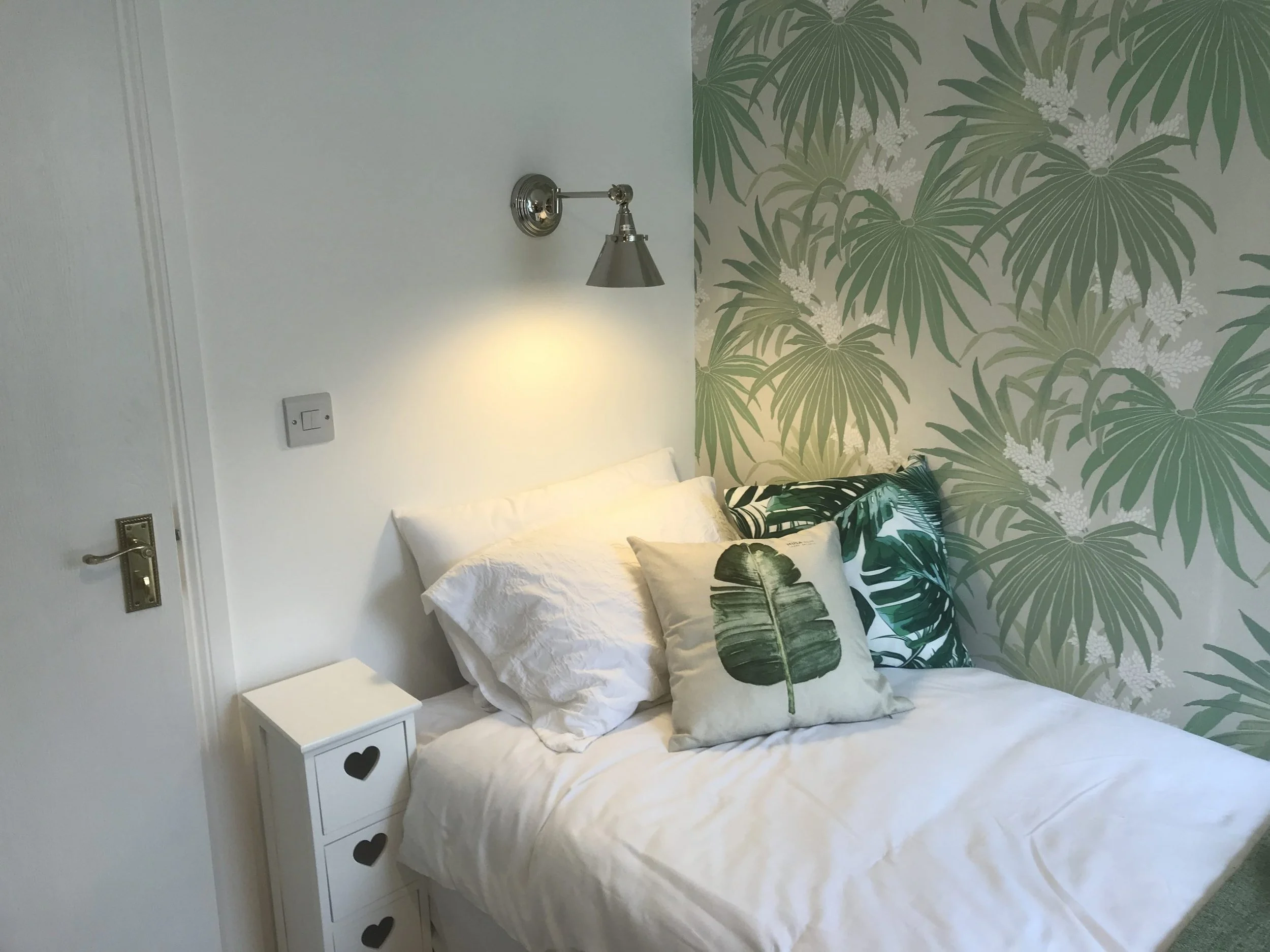 A bedroom with white walls, a bed with white bedding, and a corner accent wall decorated with green tropical leaf wallpaper. The bed has pillows with tropical leaf designs.