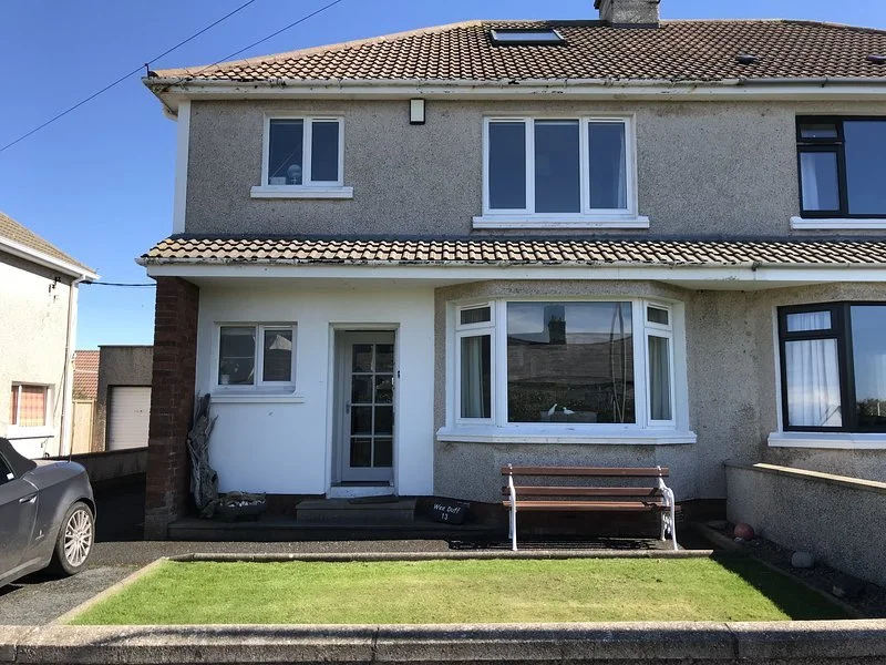 Front view of a two-story house with a beige exterior, white window frames, and a brown tiled roof. There is a small front lawn with a bench, a car parked to the left, and a clear blue sky.