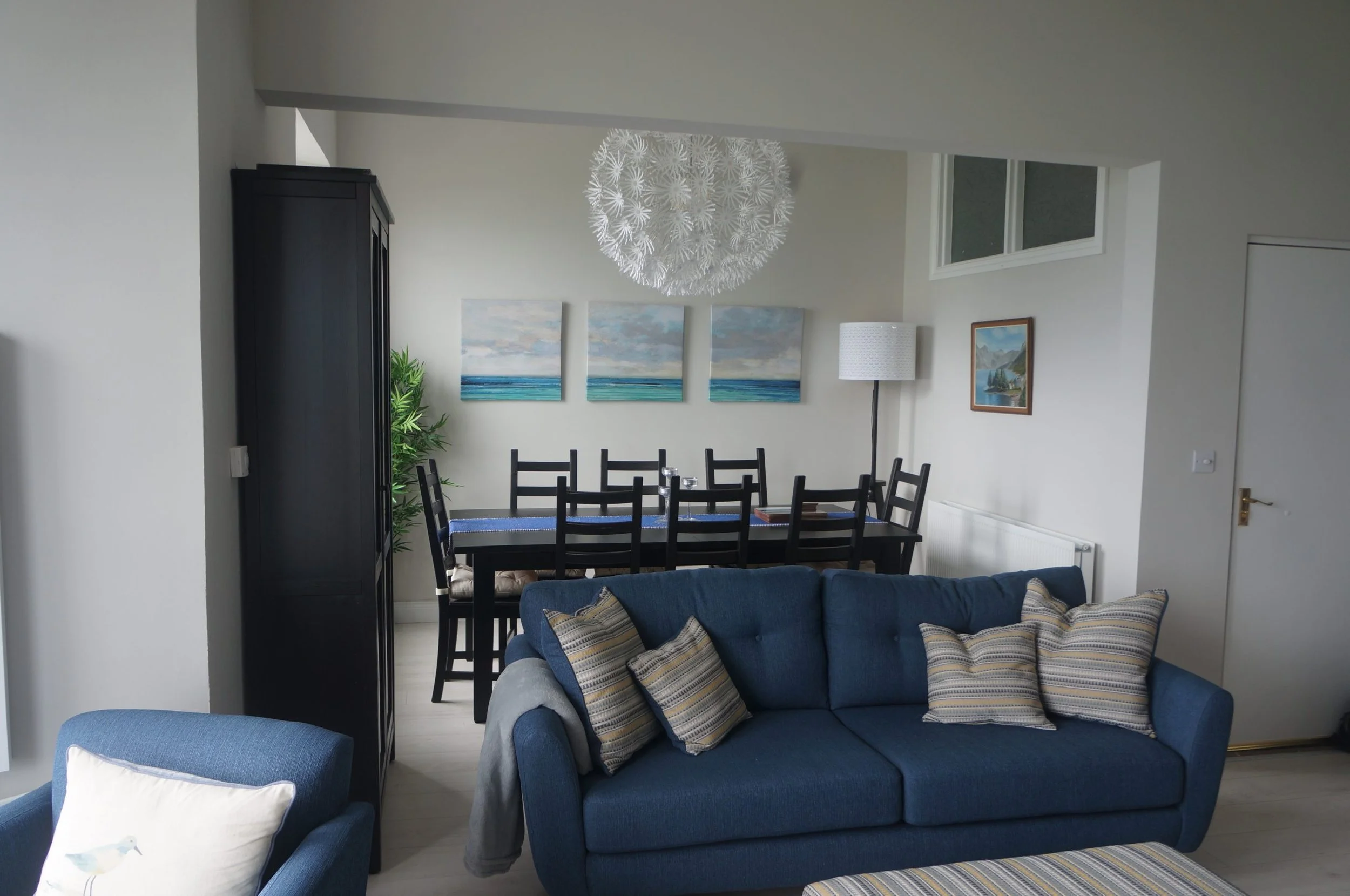 Living room with blue sofa with striped pillows, dining area with black table and chairs, artwork on walls, white textured ceiling light, and a standing lamp.