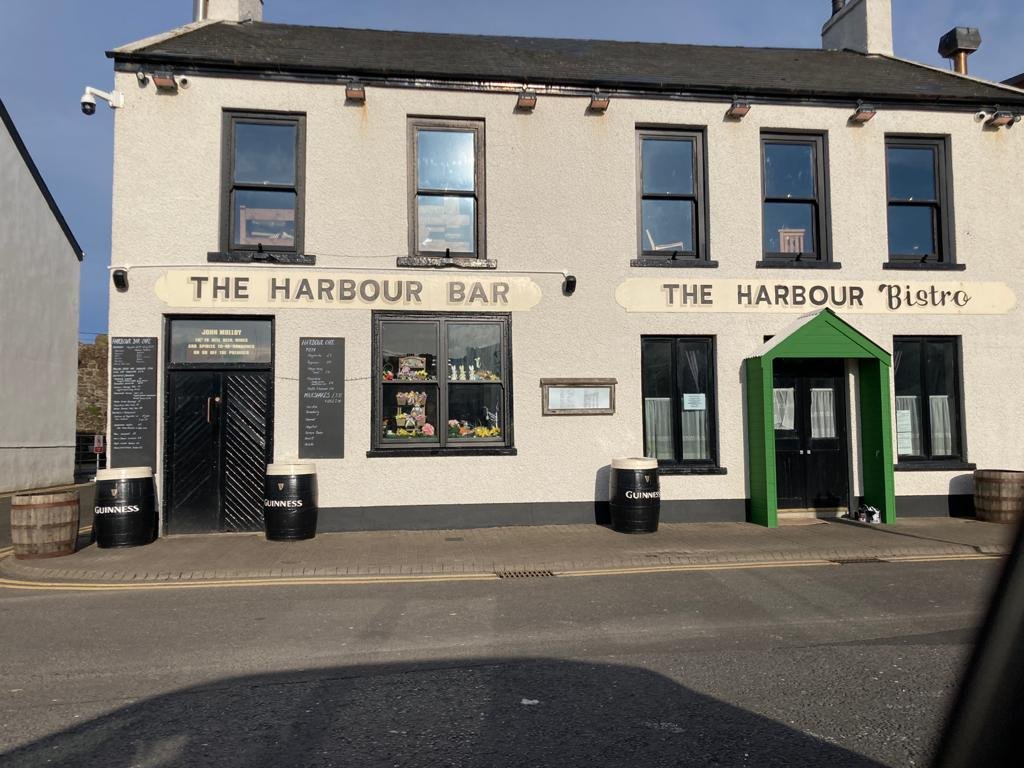 Exterior of a two-story building with signs indicating 'The Harbour Bar' and 'The Harbour Bistro'; the facade is white with black window frames, a green entrance porch, and advertising barrels outside.