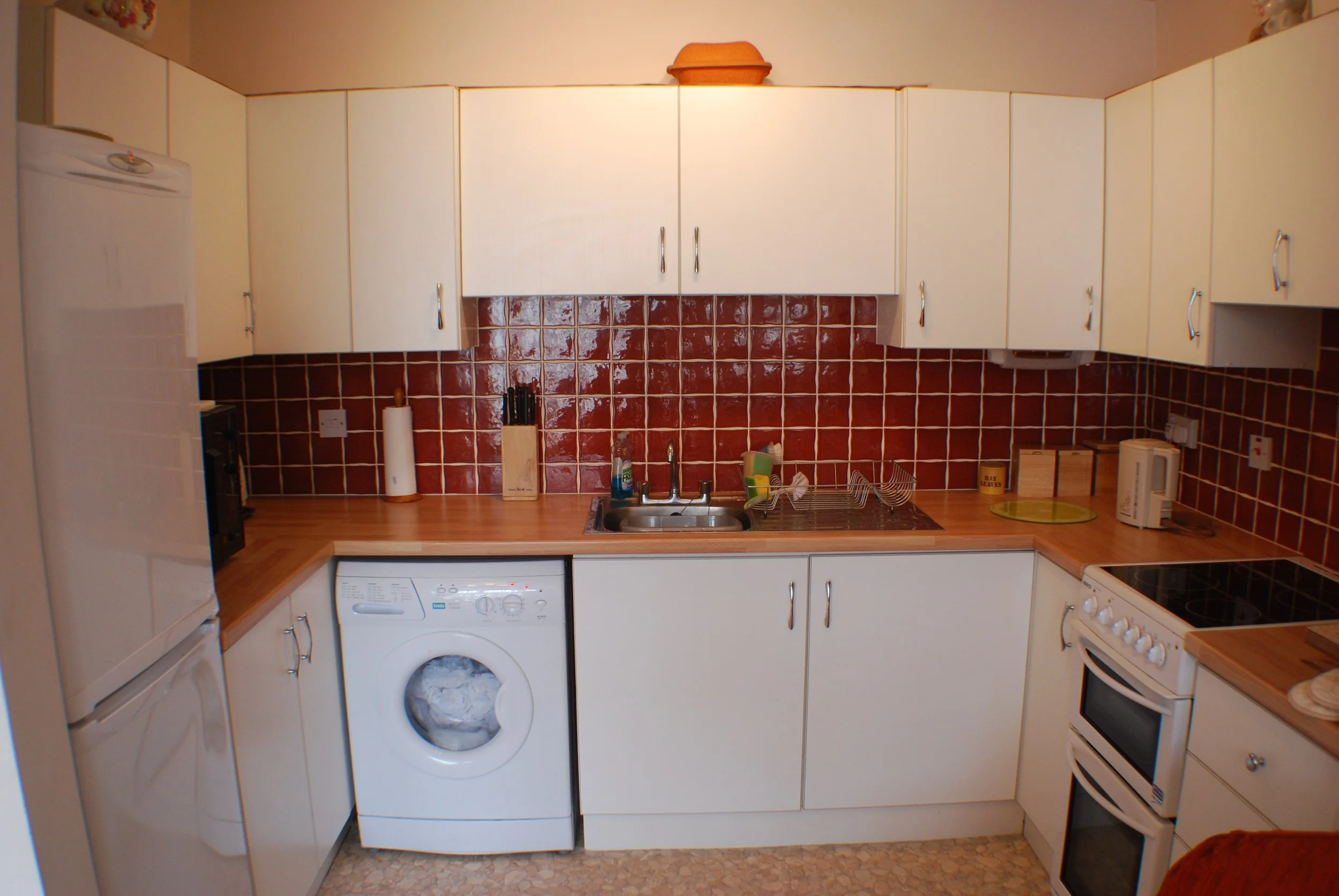 Kitchen with white cabinets, red tiled backsplash, wooden countertops, refrigerator, washing machine, stove, sink, and various small appliances and kitchen utensils.
