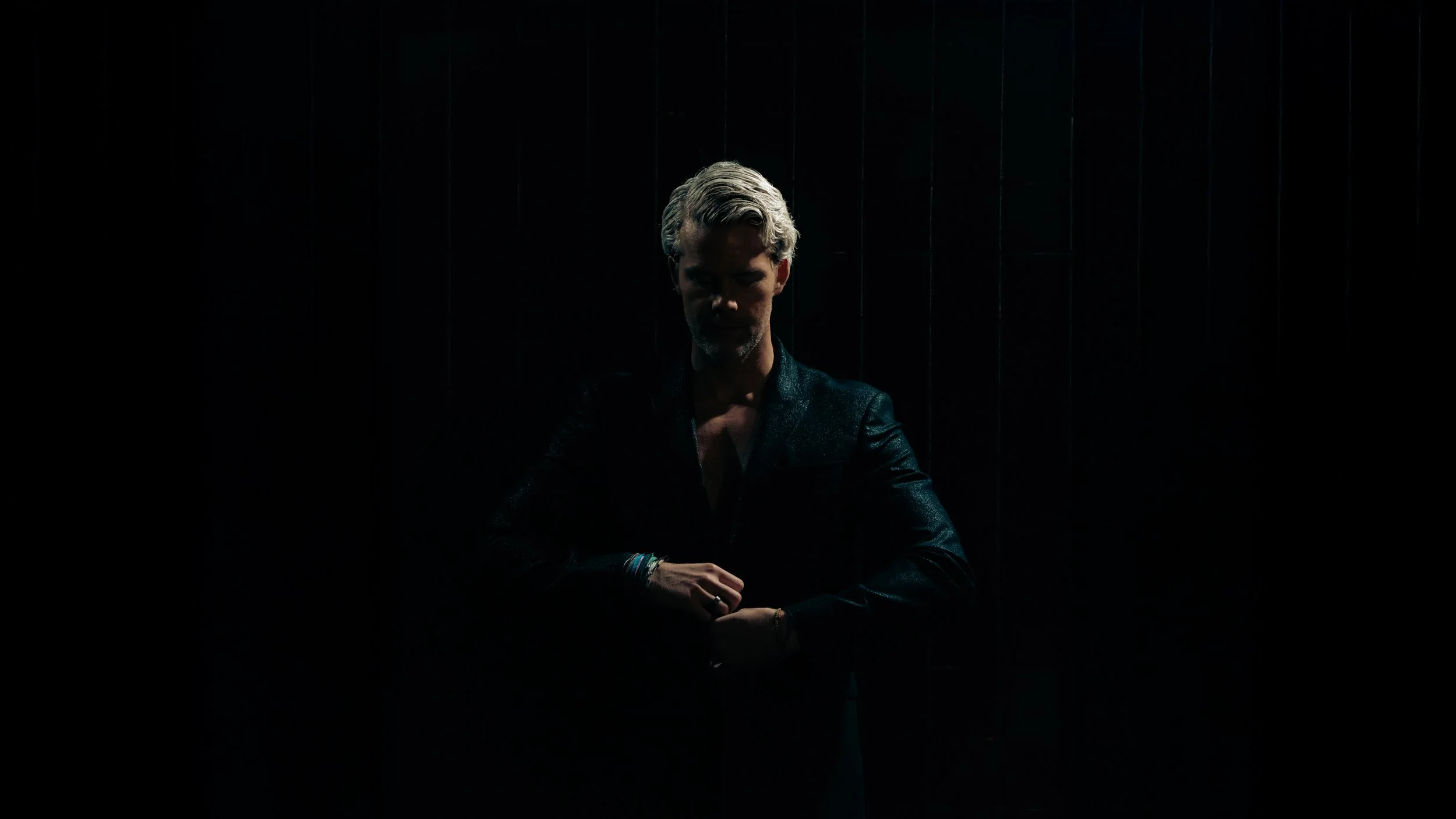 A man with light hair and a beard, dressed in a dark jacket, stands in a dimly lit space with a dark background, with low lighting focusing on his face and hands.