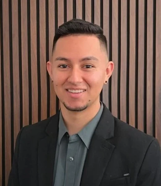 Young man wearing a dark suit and gray shirt, standing in front of a wooden-paneled wall, smiling.
