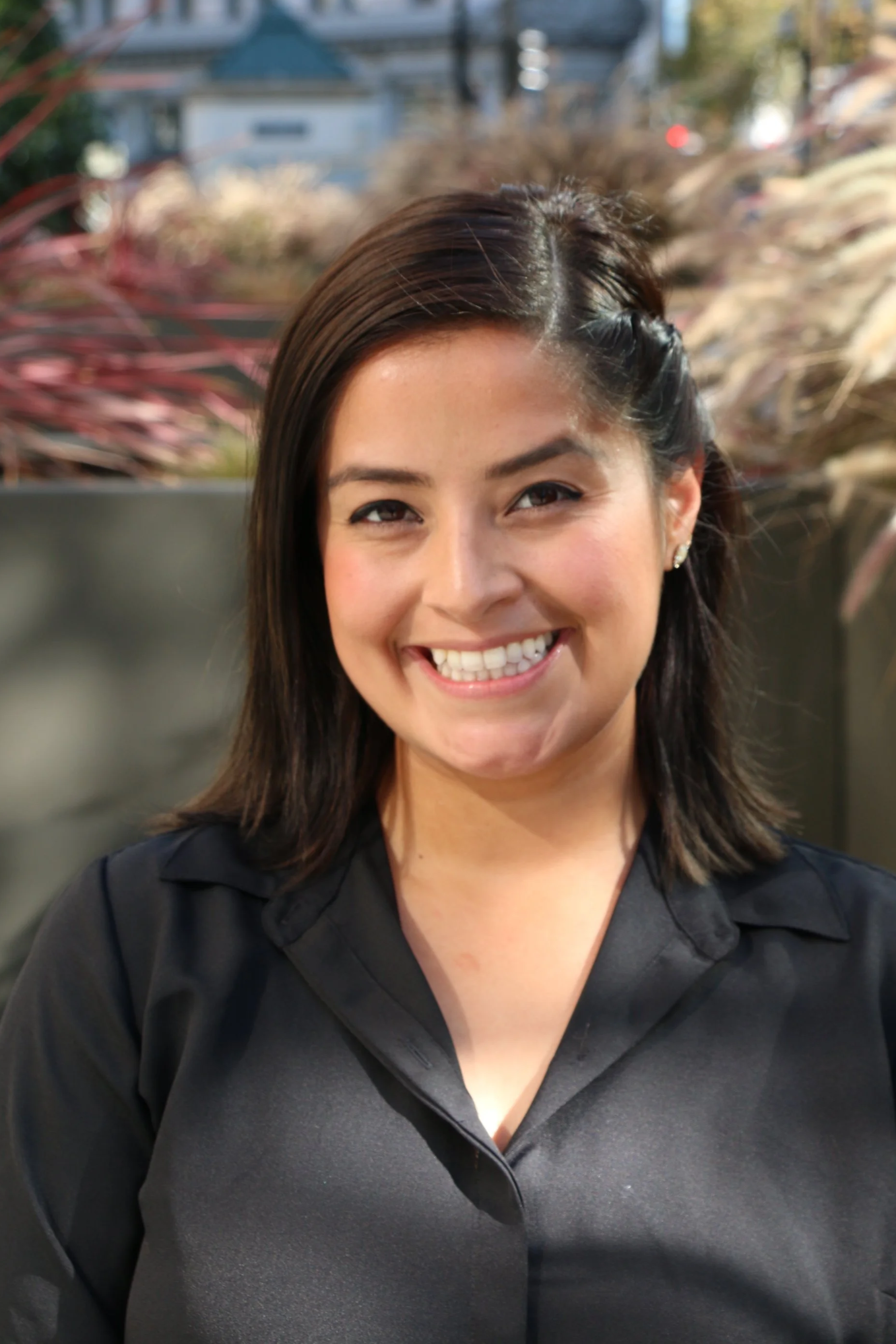 Close-up of a woman smiling outdoors, with plant foliage and houses in the background.