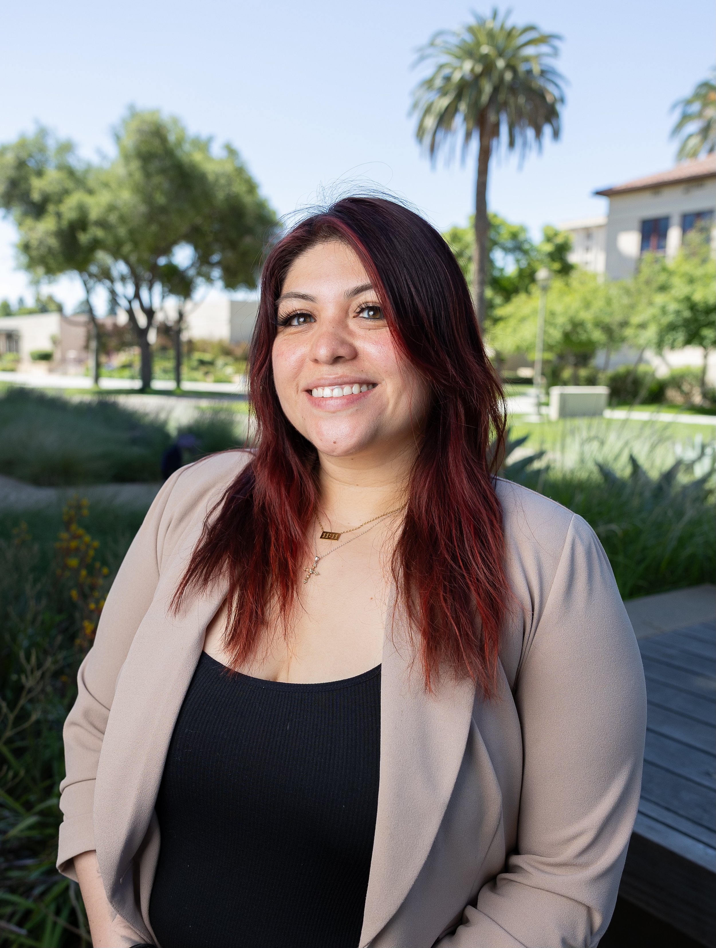 A woman with long dark red hair, wearing a beige blazer over a black top, smiling outdoors with trees and a building in the background on a sunny day.