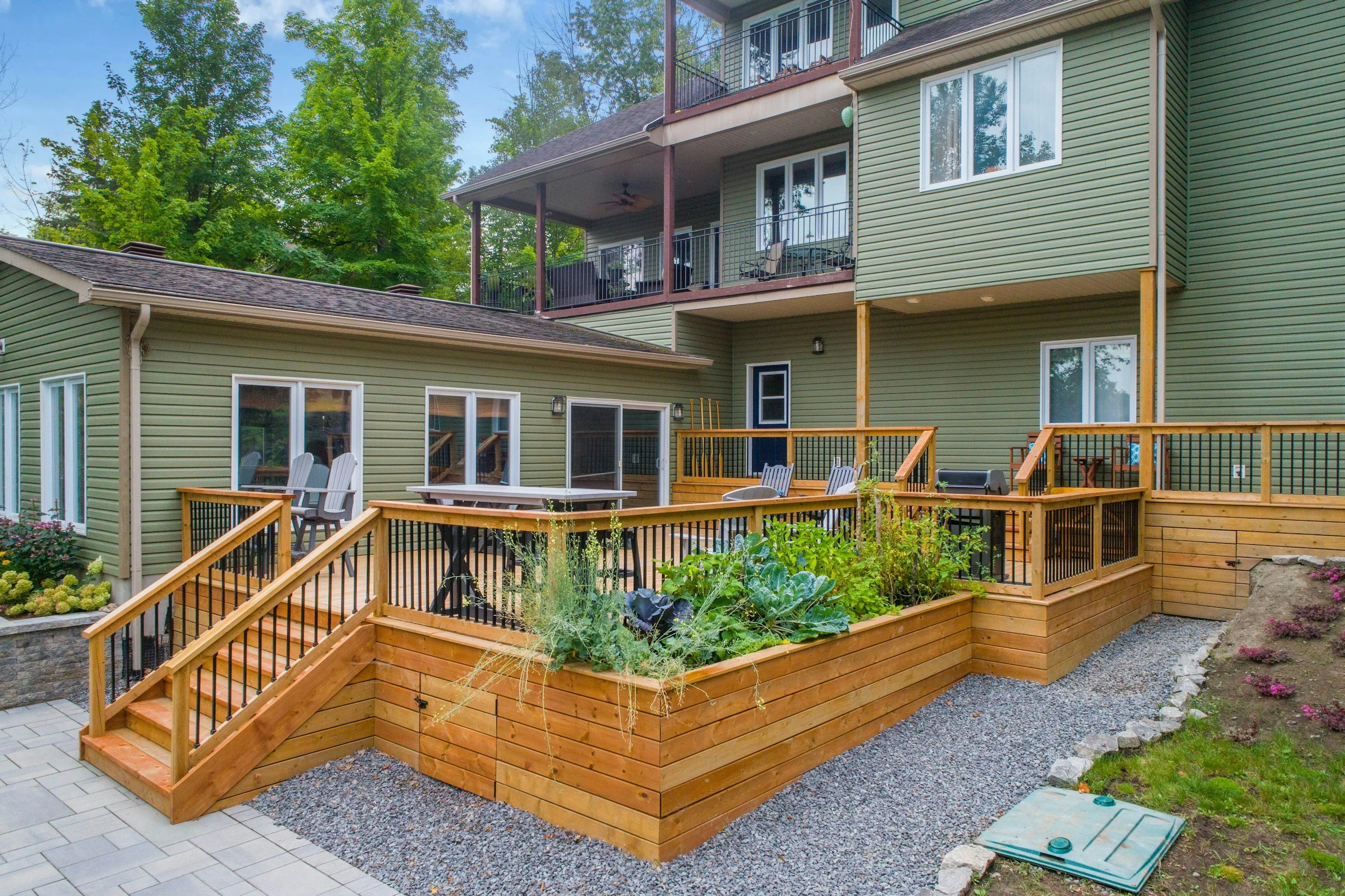 Backyard with a raised wooden deck featuring outdoor seating and grilling area, attached to a green multi-story house with sliding glass doors and large windows, surrounded by trees and landscaping.