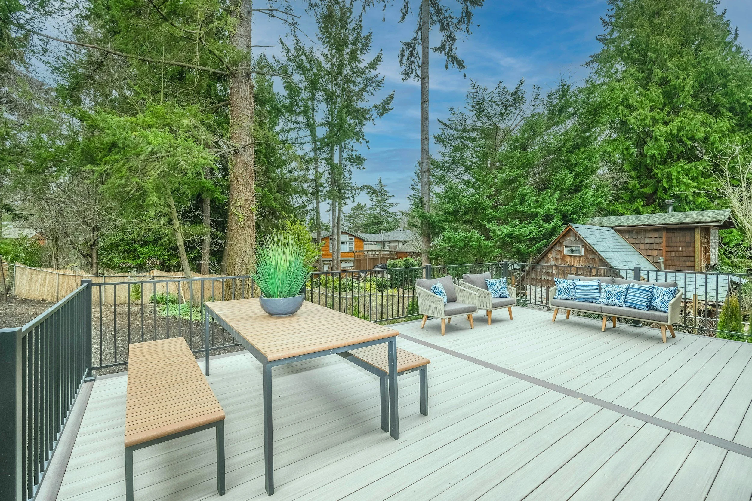 A spacious outdoor deck with light-colored wooden flooring, surrounded by a black fence, furnished with a wooden table, benches, and cushioned chairs, overlooking a lush green backyard with tall trees and a small wooden shed