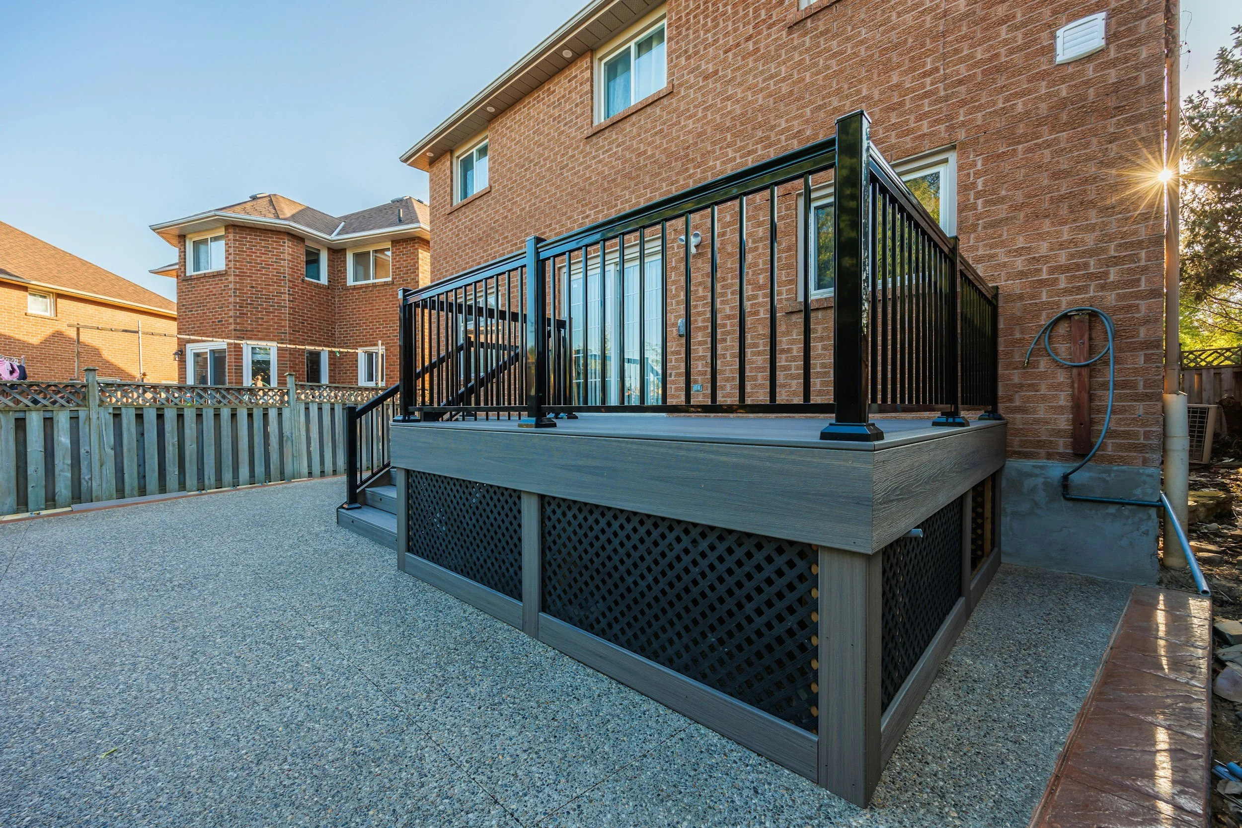 Backyard patio view of a raised wooden deck with black metal railing attached to a brick house, with neighboring brick houses in the background, evening sunlight, and a clear sky.