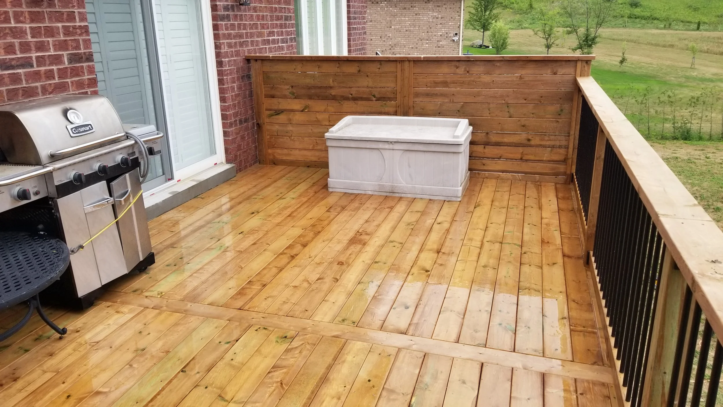 A wooden deck with a stainless steel gas grill, a white storage chest, and a wooden fence, overlooking a grassy yard with small trees.