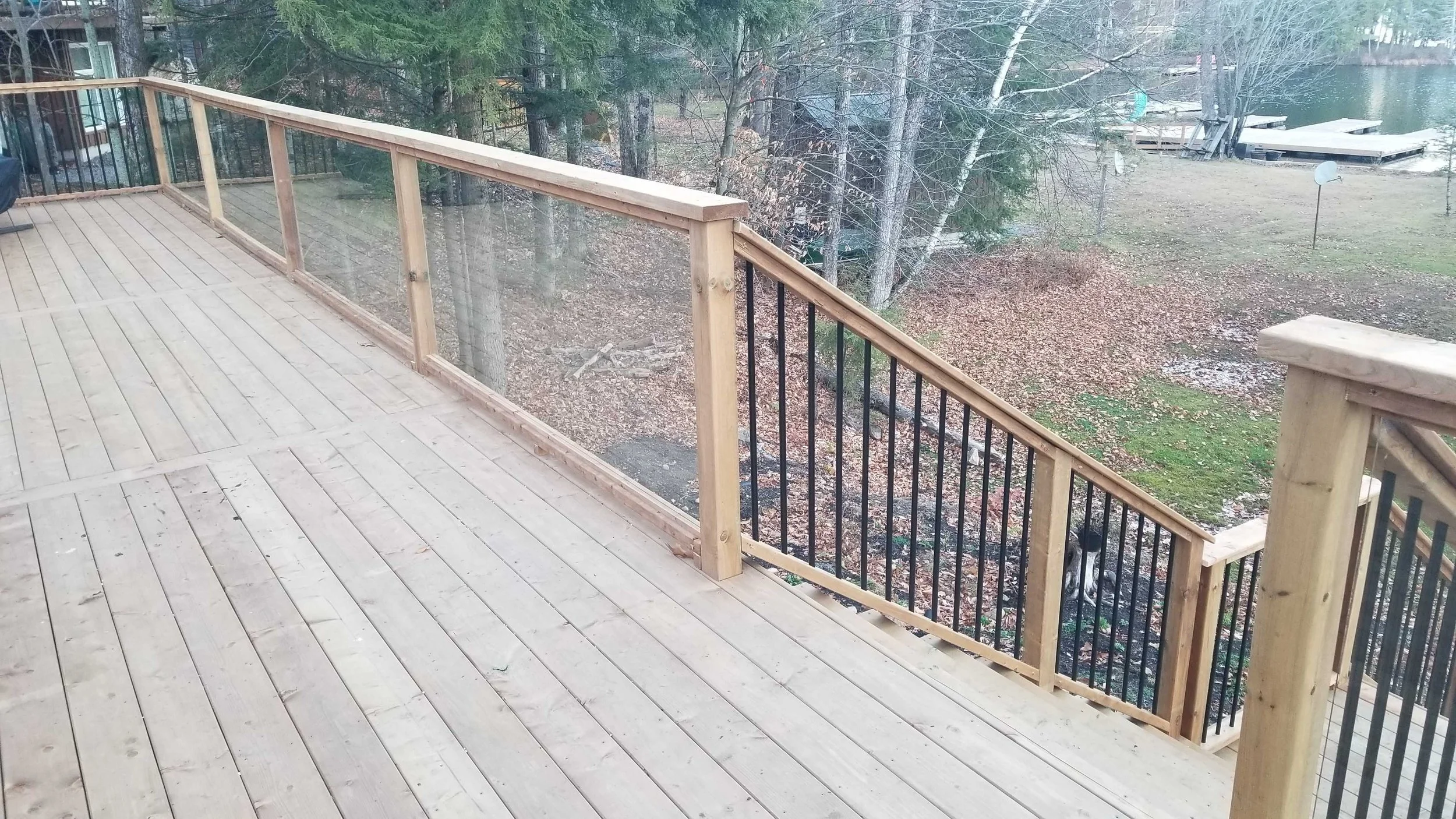 Newly constructed wooden deck with railing overlooking a yard and water, with trees and a boat dock in the background.