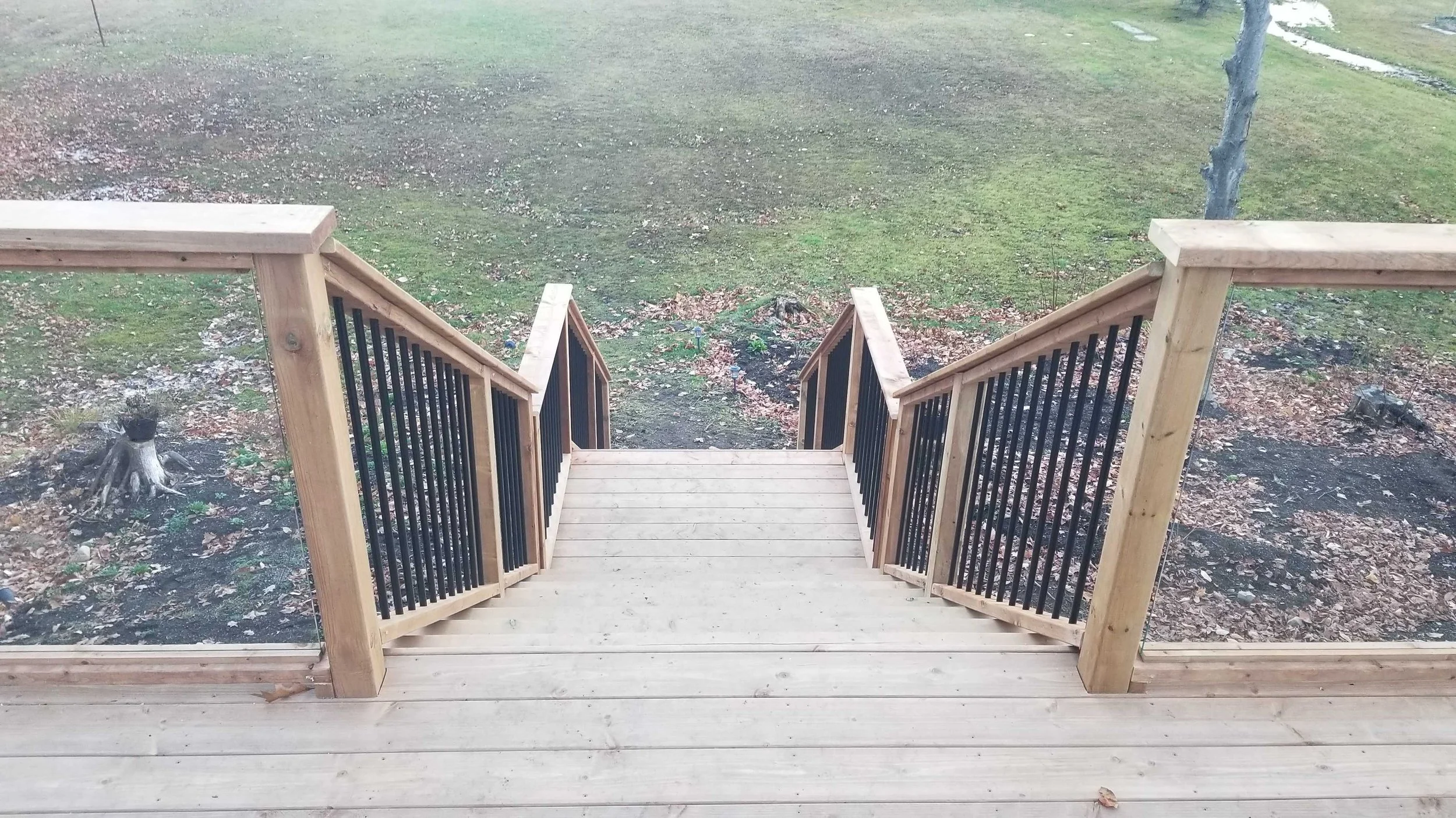 A newly built wooden deck with stairs leading down to a backyard lawn and garden area, surrounded by trees.