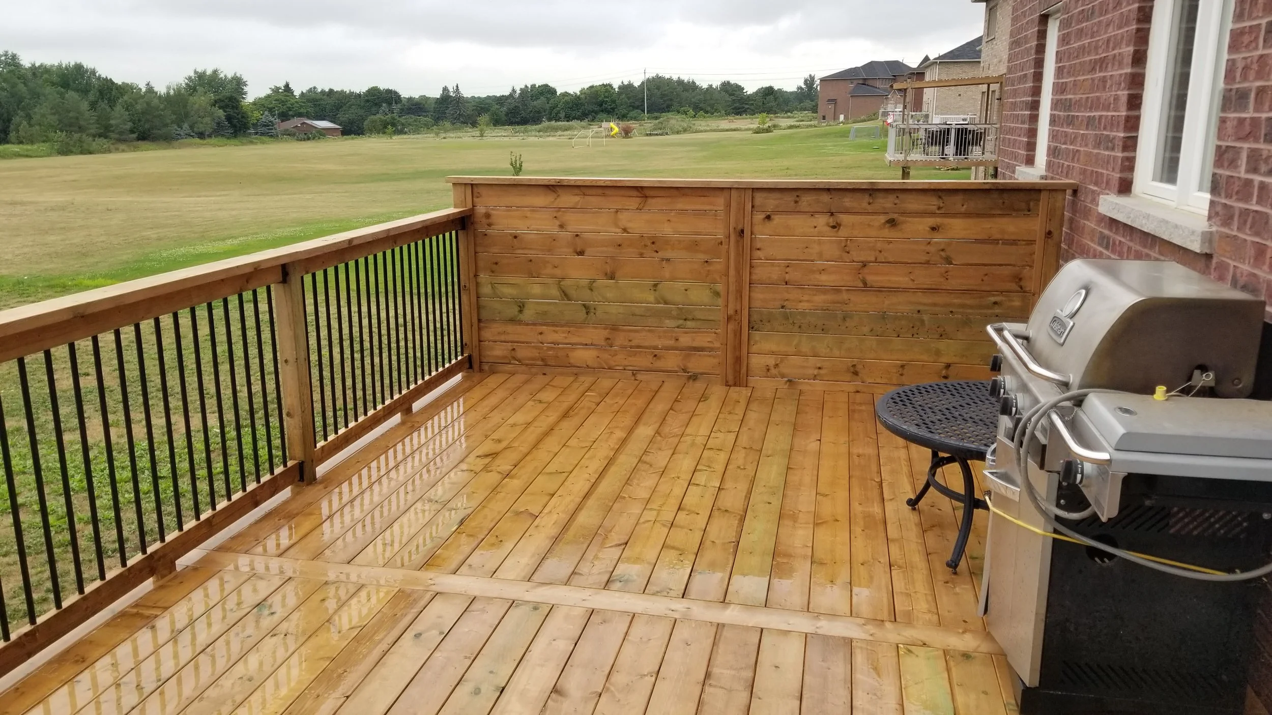 Photo of a newly built wooden deck with a black metal railing outside a brick house with a barbecue grill and a small black metal table. In the background, there is an open grassy field with some trees and a few houses visible in the distance.