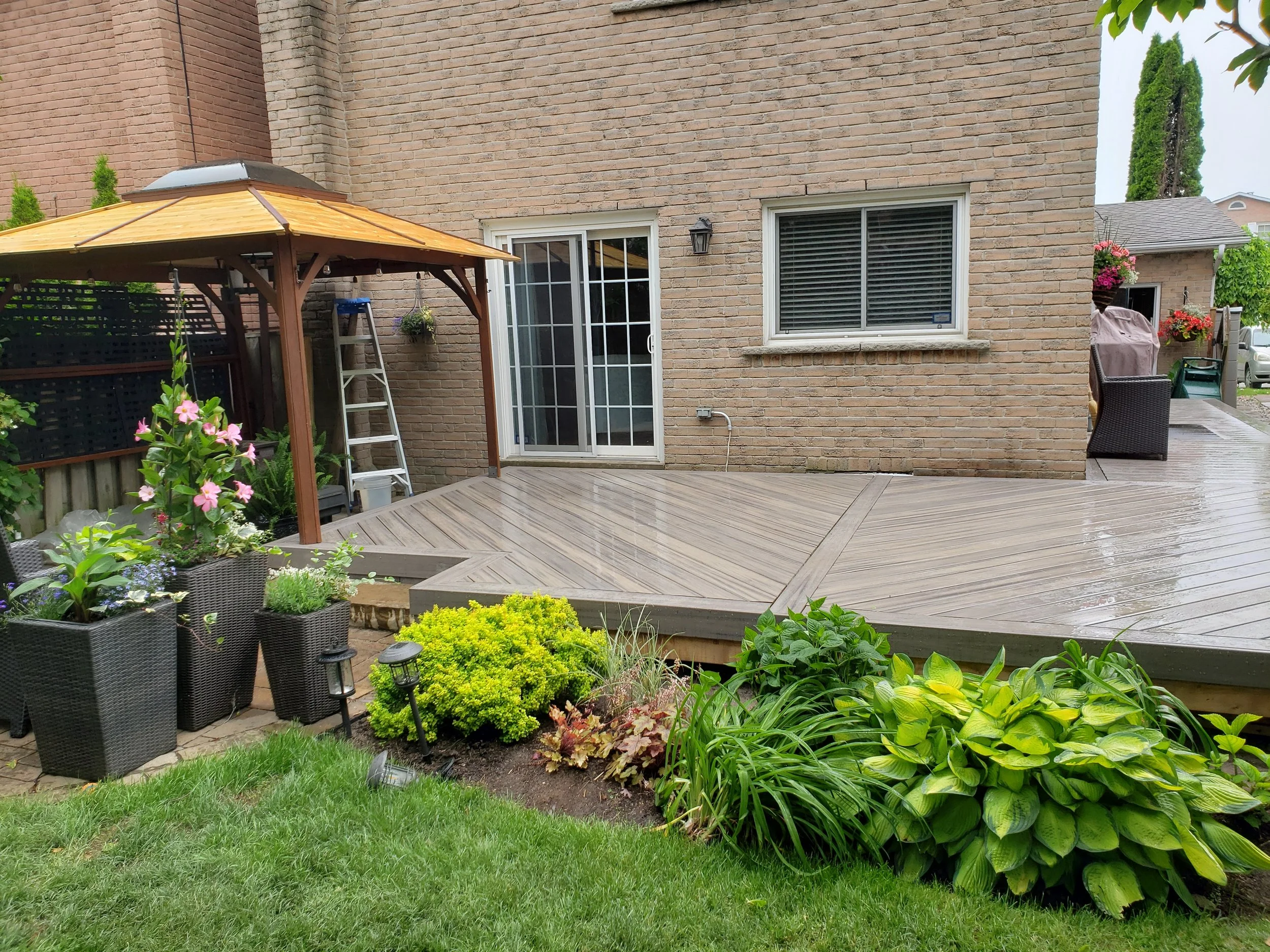 Backyard with wooden deck, colorful garden plants, a patio umbrella, ladder, and brick house wall with sliding door and window.