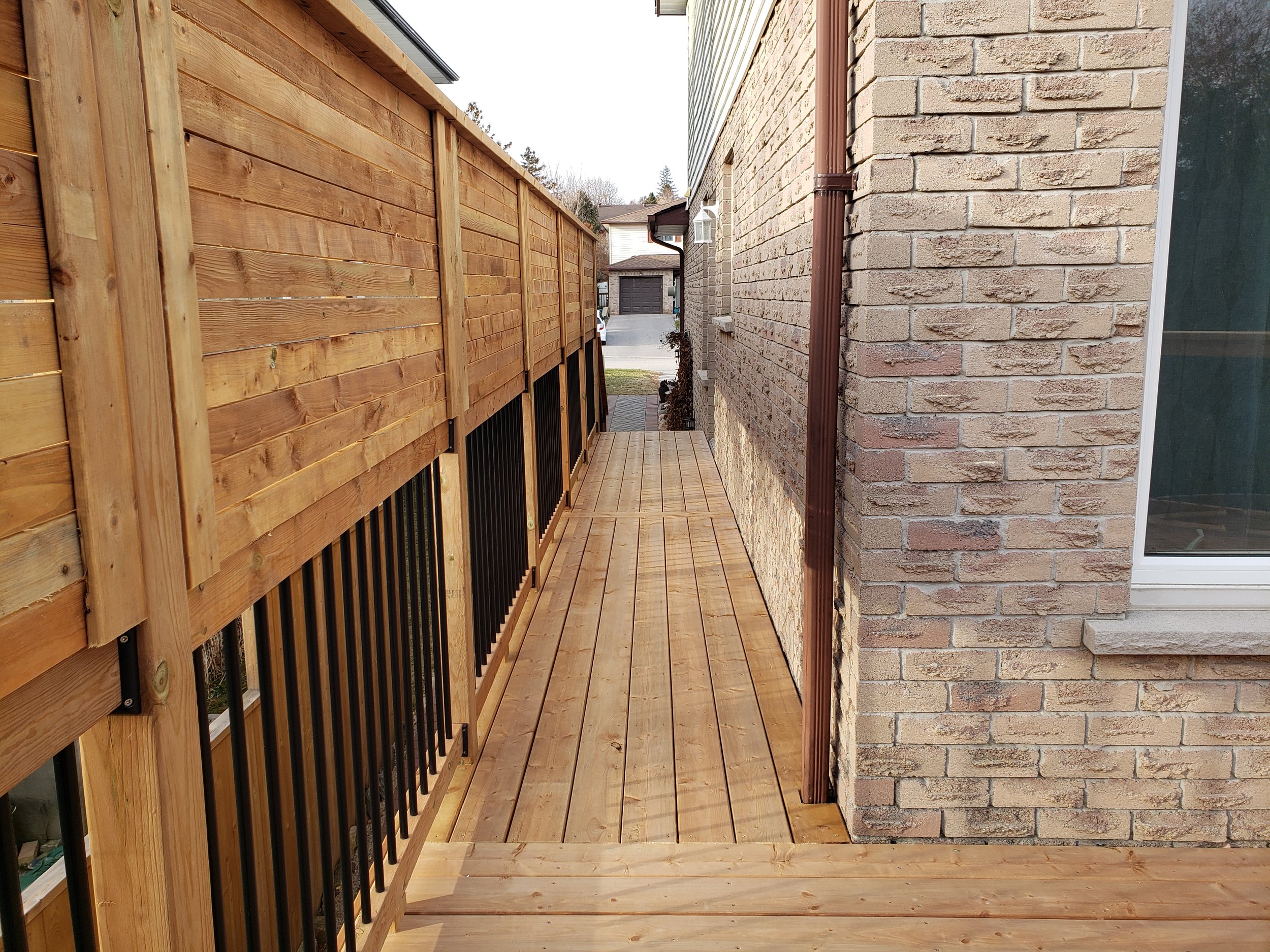 Newly built wooden deck walkway alongside a brick house wall, with black metal railing on the left side and a window on the right.