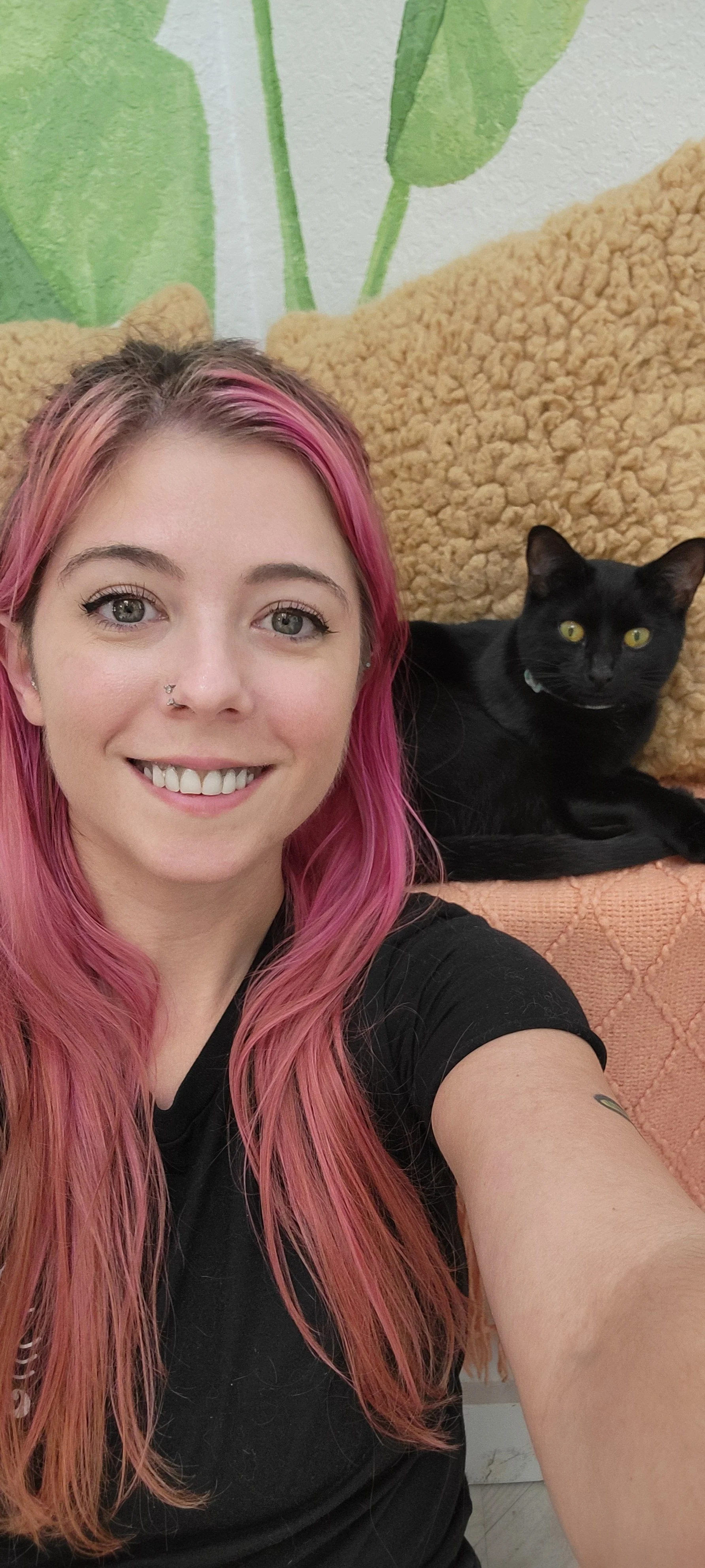 A young woman with pink hair and a septum piercing taking a selfie with her black cat on an orange and beige couch, with a green leafy mural in the background.