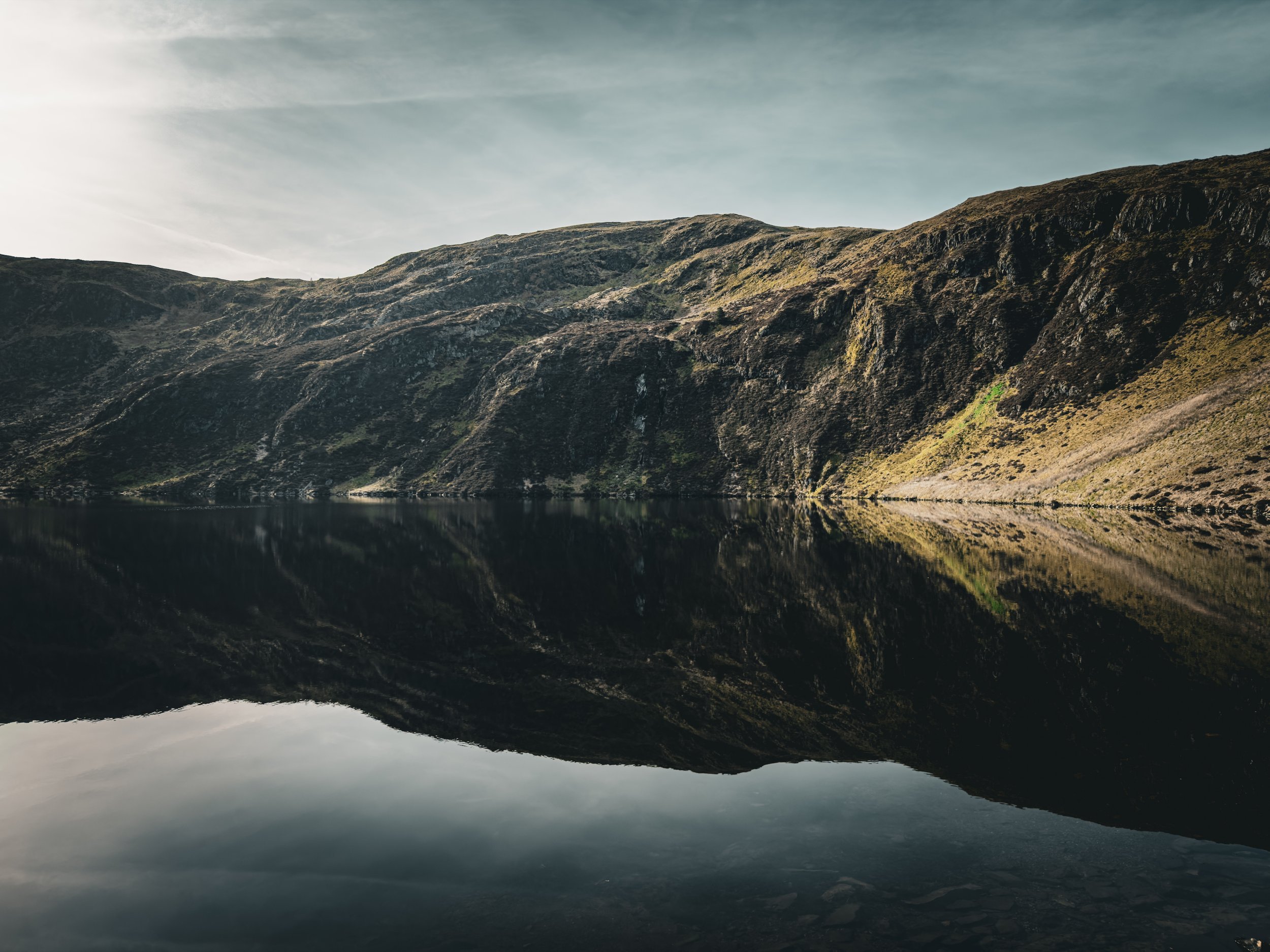 A scenic view of a lake surrounded by steep, green, moss-covered hills with their reflection visible in the calm water, under a partly cloudy sky.